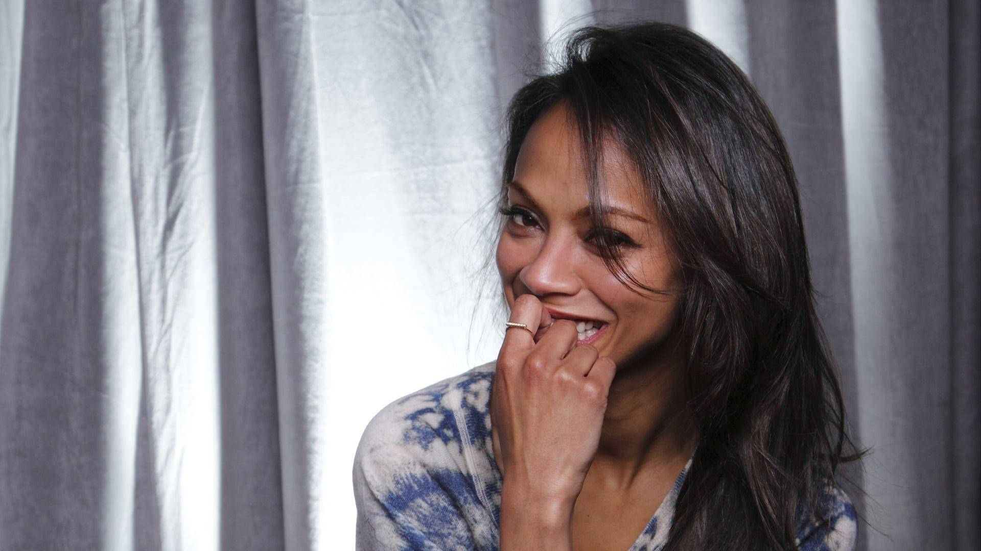 Zoe Saldana 桌布 壁紙 — 一位深色頭髮的女性在銀色窗簾背景下咬著下唇頑皮地微笑。