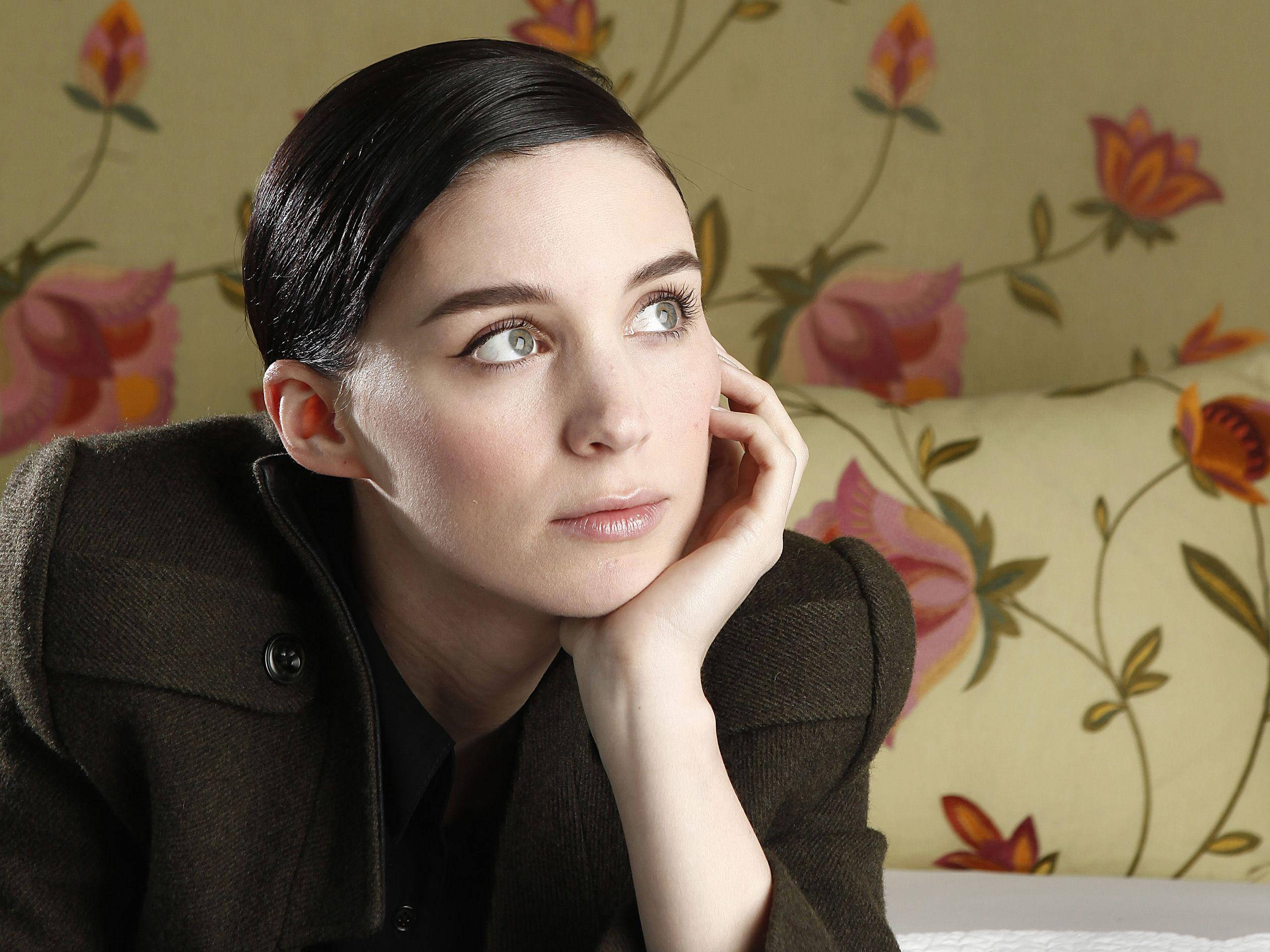 Rooney Mara 桌布 壁紙 — 一位深色頭髮的女性將下巴擱在手上，在花卉壁紙背景下若有所思地向上凝視。