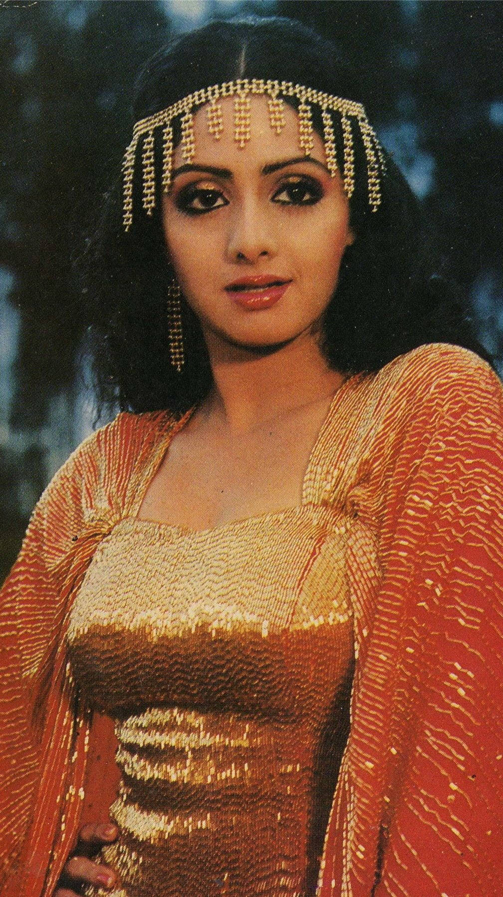 Sridevi 桌布 壁紙 — 一位深色頭髮的女性戴著金色珠飾頭飾，身穿閃閃發光的橙色和金色亮片服裝，背景是模糊的森林。