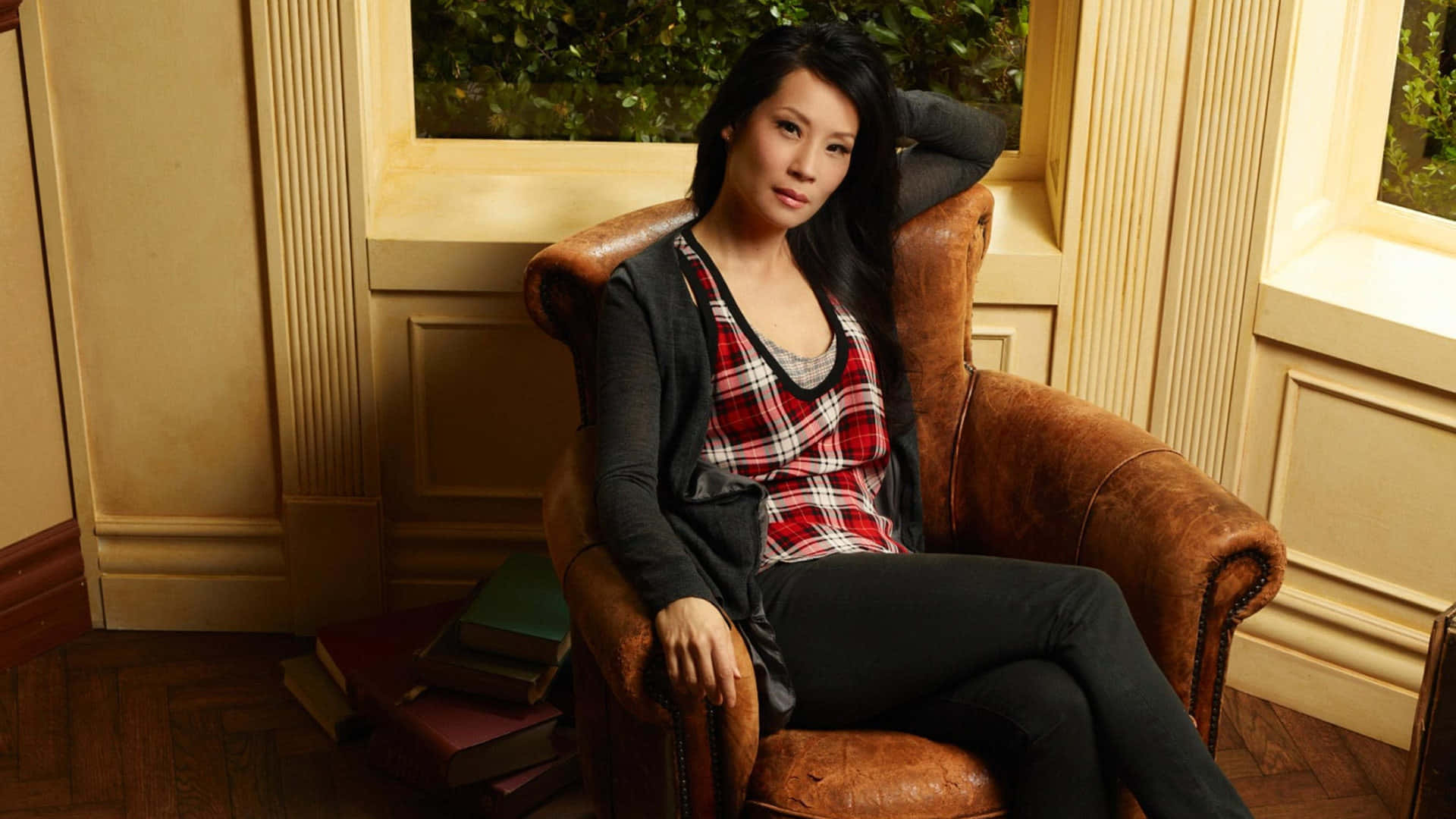 lucy liu 桌布 壁紙 — 一位深色頭髮的女性放鬆地坐在窗邊的復古皮革扶手椅上，附近堆疊著書籍。