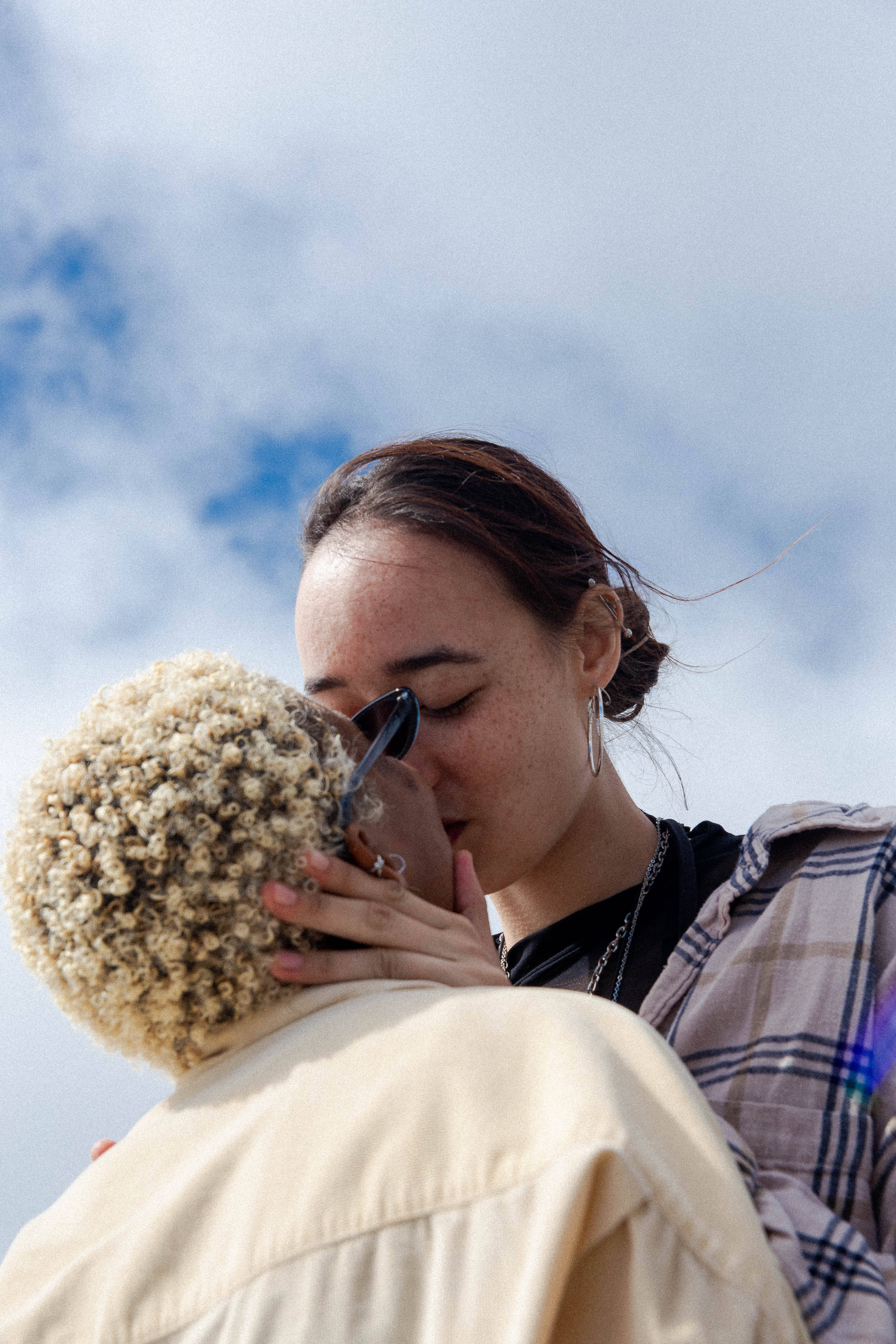 女同志 桌布 壁紙 — 一位深色頭髮的女性正在與另一位捲曲金髮的人接吻，背景是明亮的藍天和散落的雲朵。