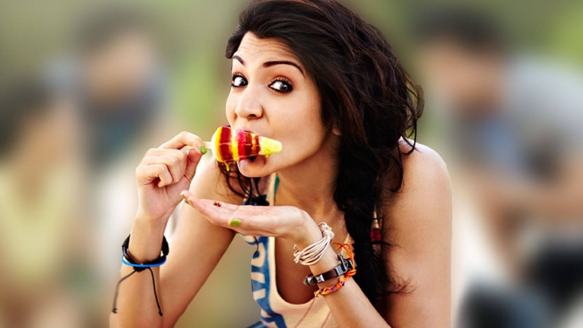 Anushka Sharma 桌布 壁紙 — 一位深色頭髮的年輕女子，一邊用一隻手拿著色彩繽紛的條紋冰棒，一邊用另一隻手掌接住滴落的汁液，俏皮地咬了一口。