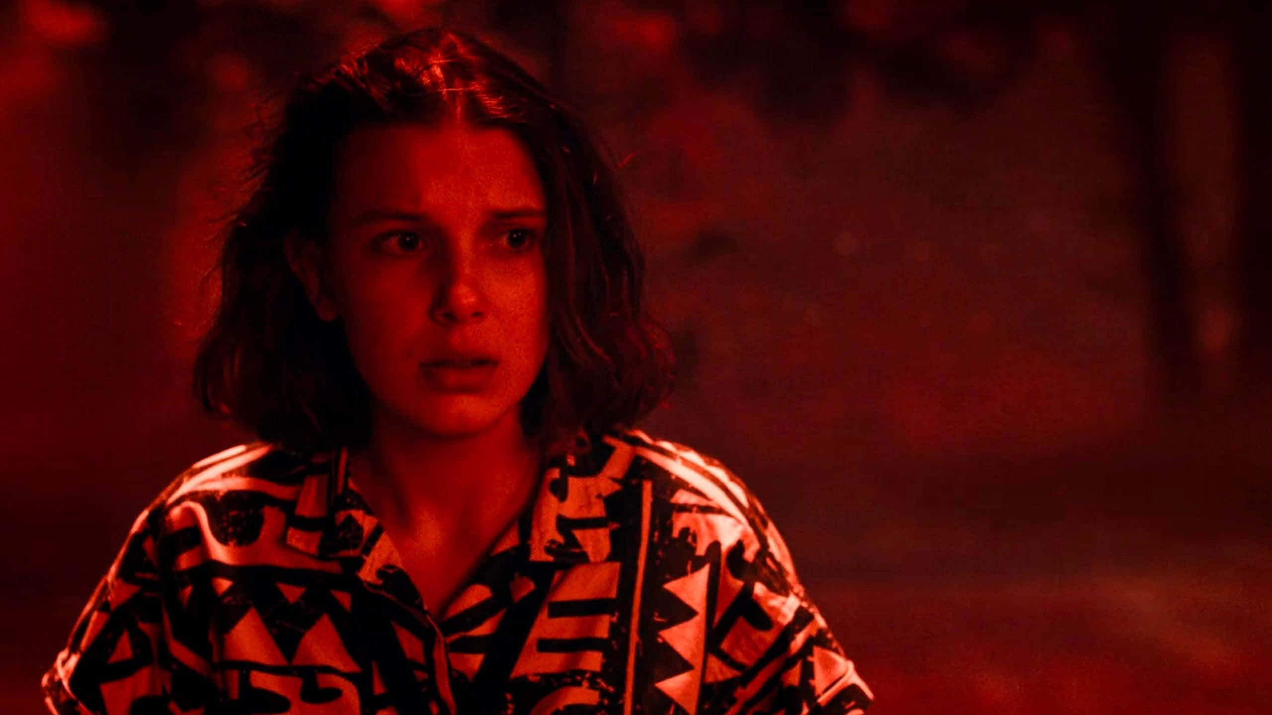 Eleven Stranger Things 桌布 壁紙 — 一位深色頭髮的年輕女子在不祥紅光照耀的昏暗場景中凝視著。