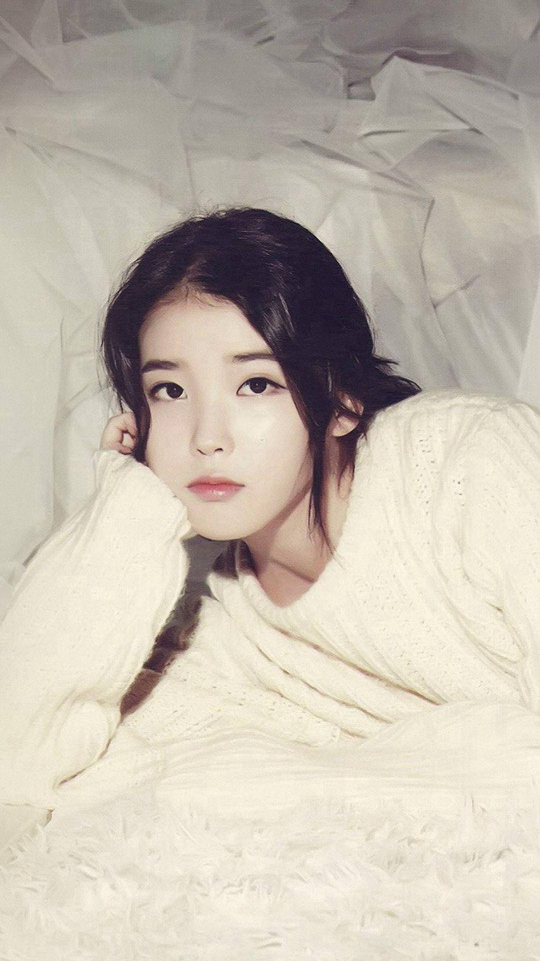 iu phone 桌布 壁紙 — 一位深色頭髮的年輕女子將頭靠在手上，躺在柔軟的白色床單上，穿著舒適的奶油色毛衣。