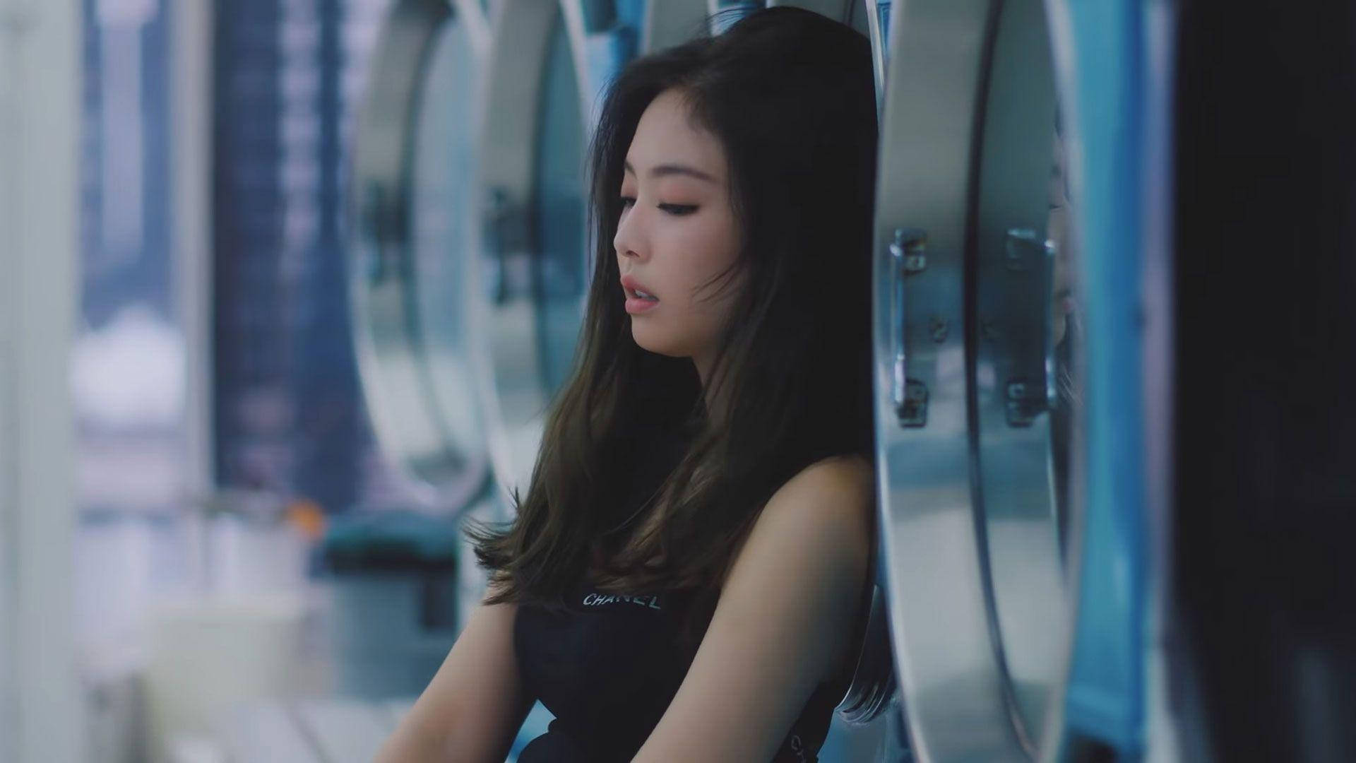 Jennie 桌布 壁紙 — 一位深色頭髮的年輕女子靠在一排工業洗衣機上，身後窗外可見模糊的城市建築。