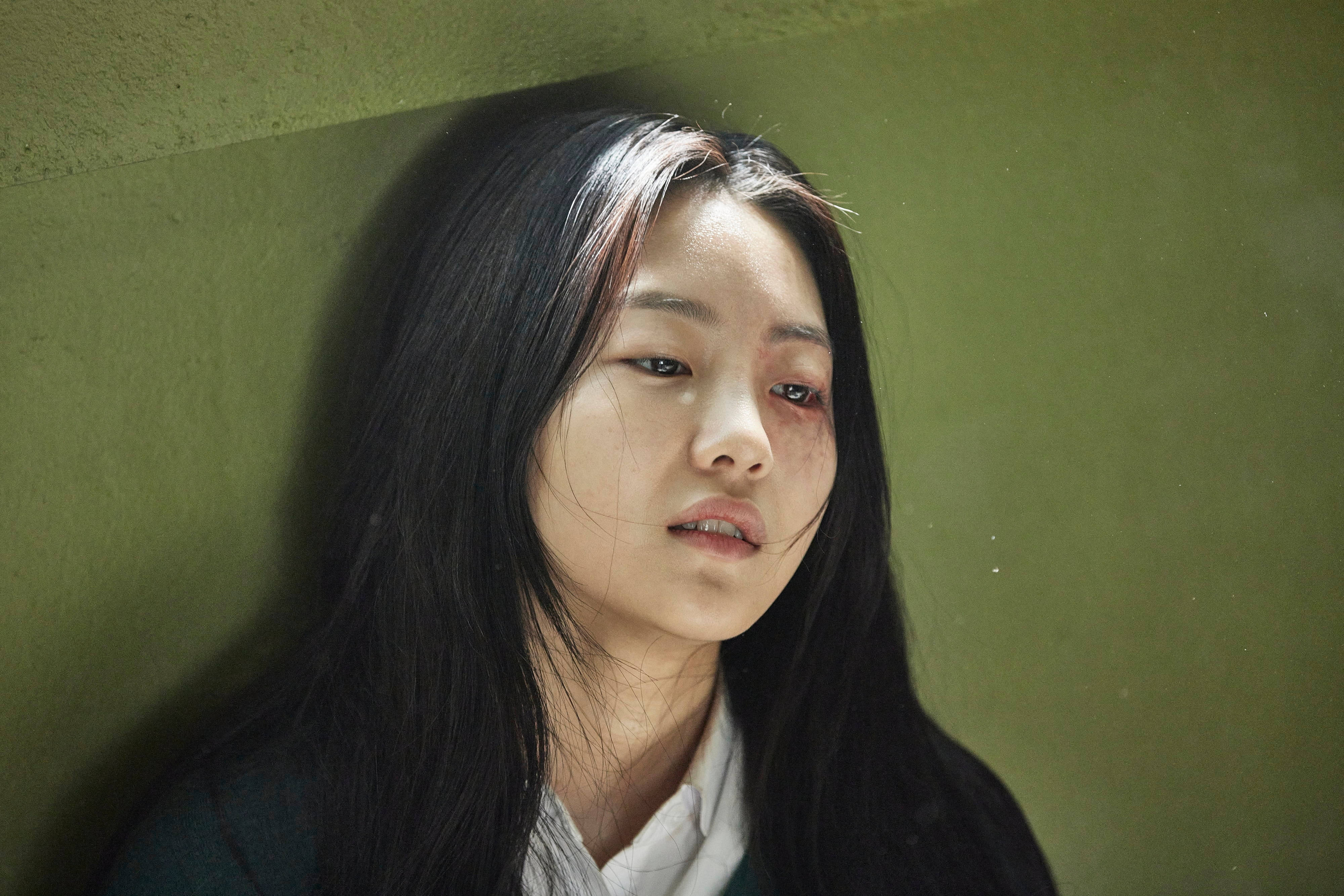 殭屍動圖 桌布 壁紙 — 一位深色頭髮的年輕女子靠在綠色牆上，瘀傷的眼睛和疲憊的表情在昏暗燈光下傳達出痛苦。