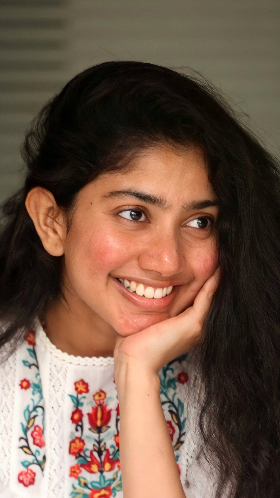 Sai Pallavi 桌布 壁紙 — 一位深色頭髮的年輕女性，在柔和模糊的背景前，將下巴擱在手上，露出溫暖的笑容。
