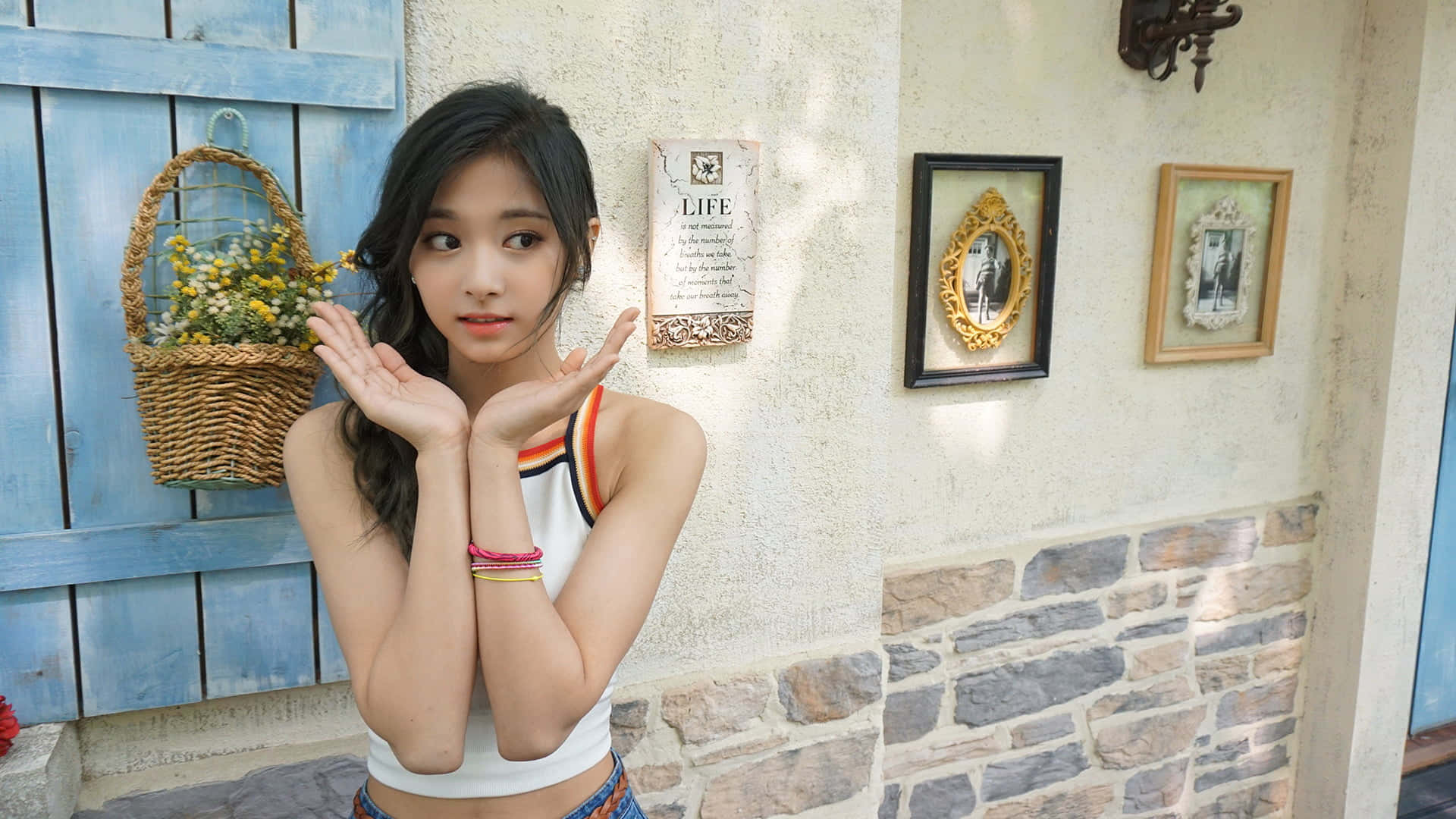 twice 桌布 壁紙 — 一位深色頭髮的年輕女性在裝飾有裱框照片和花籃的質樸石頭和藍色木牆前俏皮地擺姿勢。