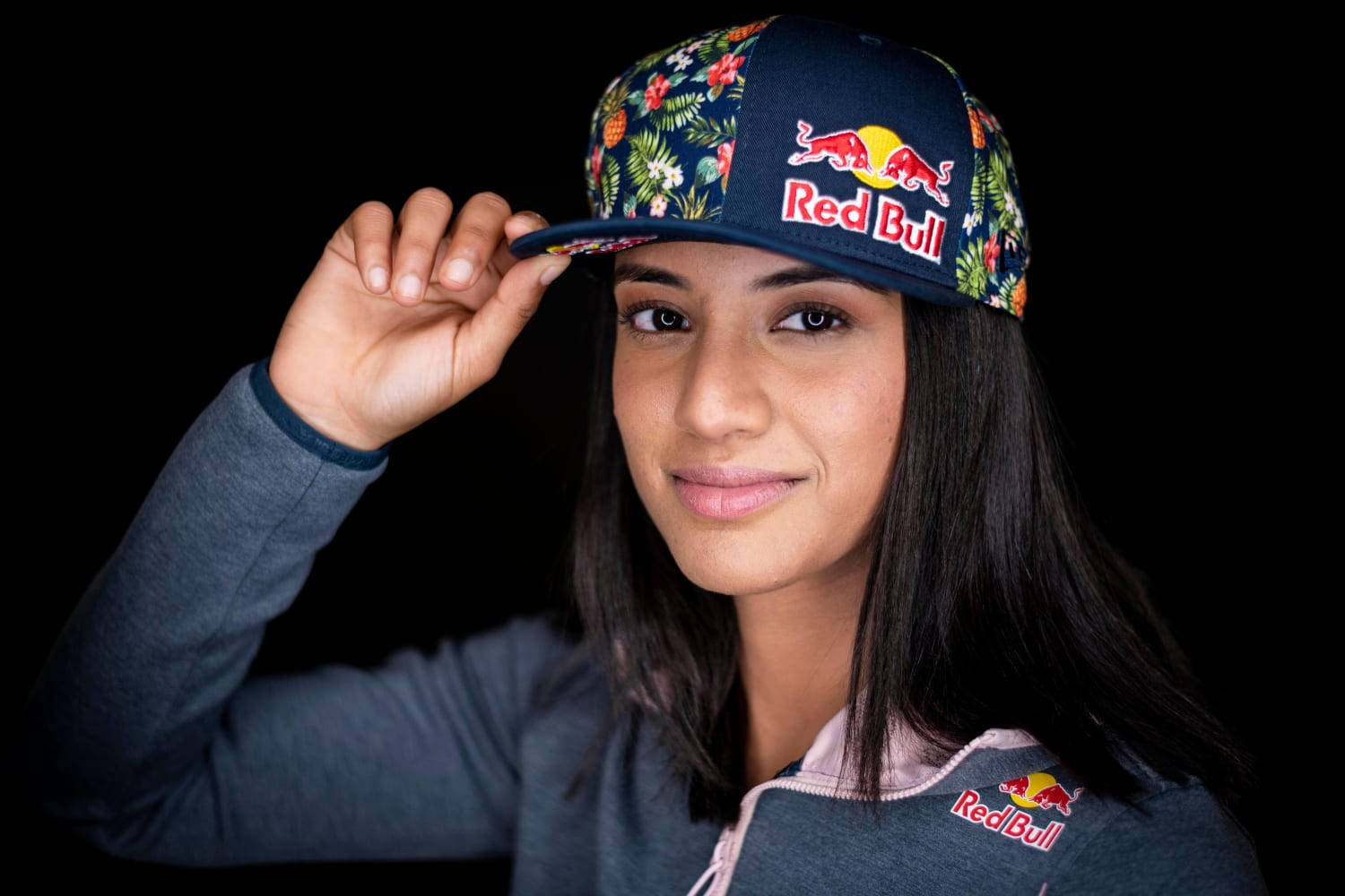 Red Bull 桌布 壁紙 — 一位深色頭髮的年輕女性在黑色背景前微笑著調整她的花卉圖案Red Bull帽子。