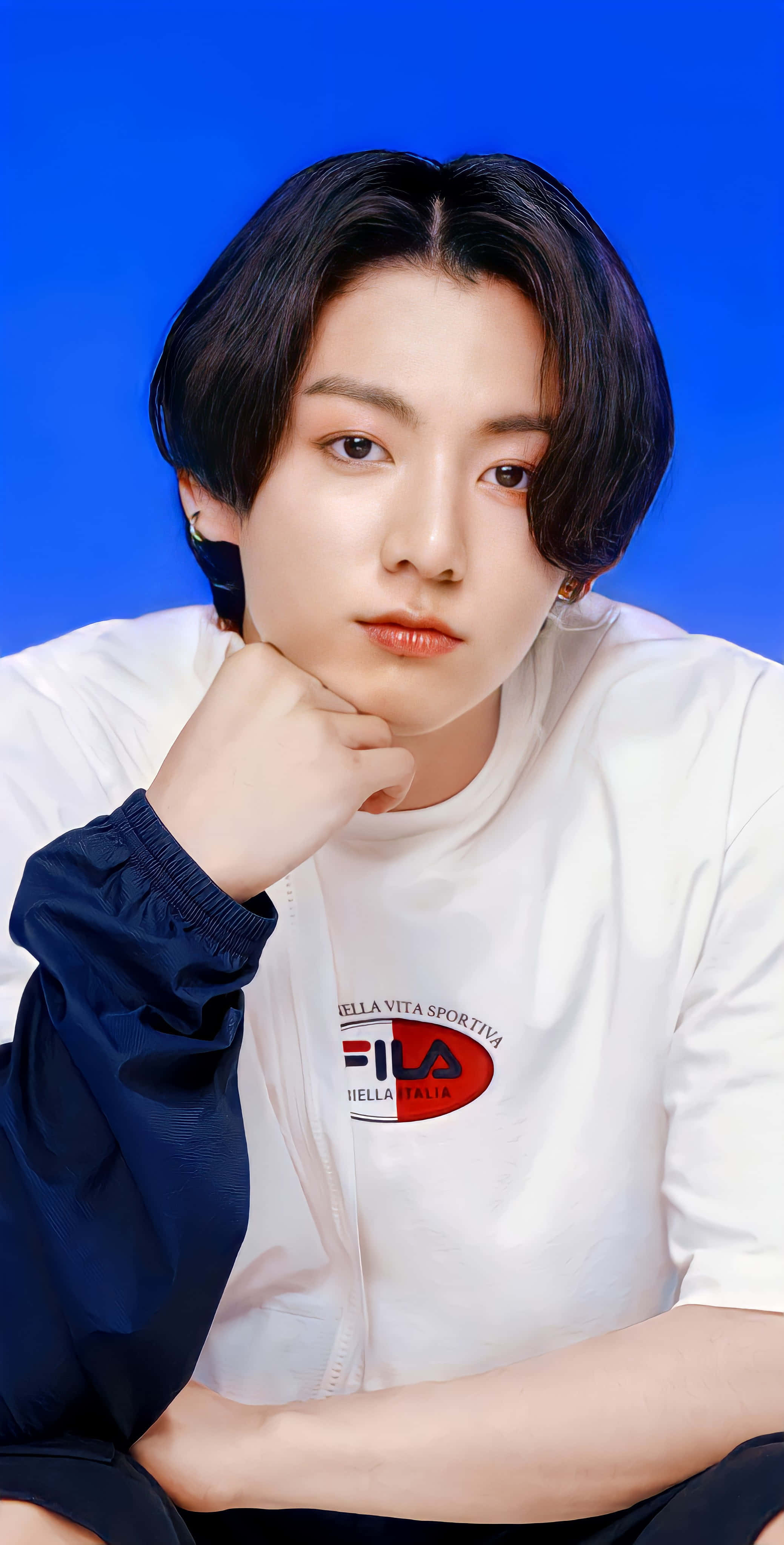 Jungkook 桌布 壁紙 — 一位深色頭髮的年輕男子，在純藍色背景前，穿著白色FILA襯衫，將下巴擱在手上。