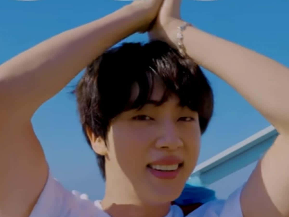 BTS 桌布 壁紙 — 一位深色頭髮的年輕男子在清澈的藍天下，雙臂高舉過頭，露出燦爛的笑容。