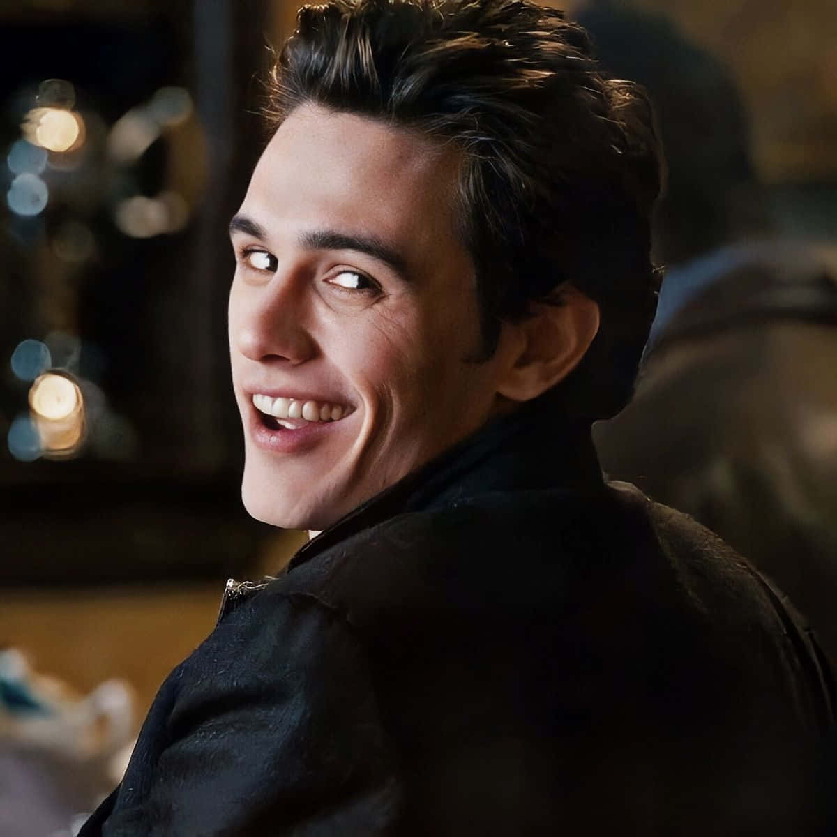 Harry Osborn 桌布 壁紙 — 一位深色頭髮的年輕男子在溫暖照明的室內環境中，身後有散景燈光，越過肩膀燦爛微笑。