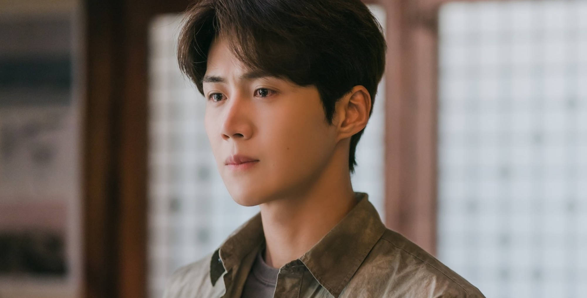 Kim Seon Ho 桌布 壁紙 — 一位深色頭髮的年輕男子在溫暖照明的室內環境中，帶著木框和柔和背景模糊，若有所思地望向一旁。