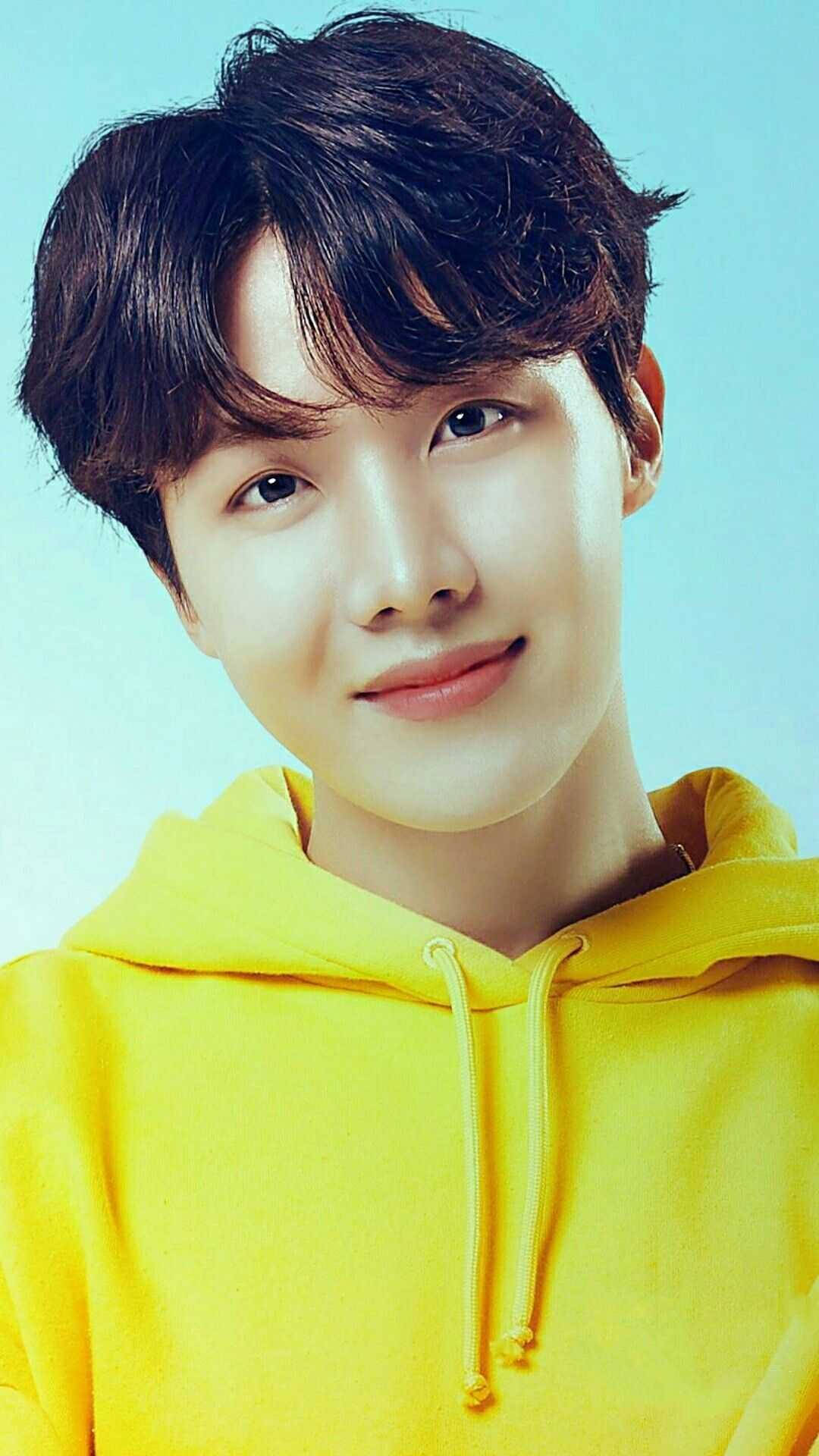 jhope 桌布 壁紙 — 一位深色頭髮的年輕男子穿著亮黃色連帽衫，在淺藍色背景前溫柔微笑。