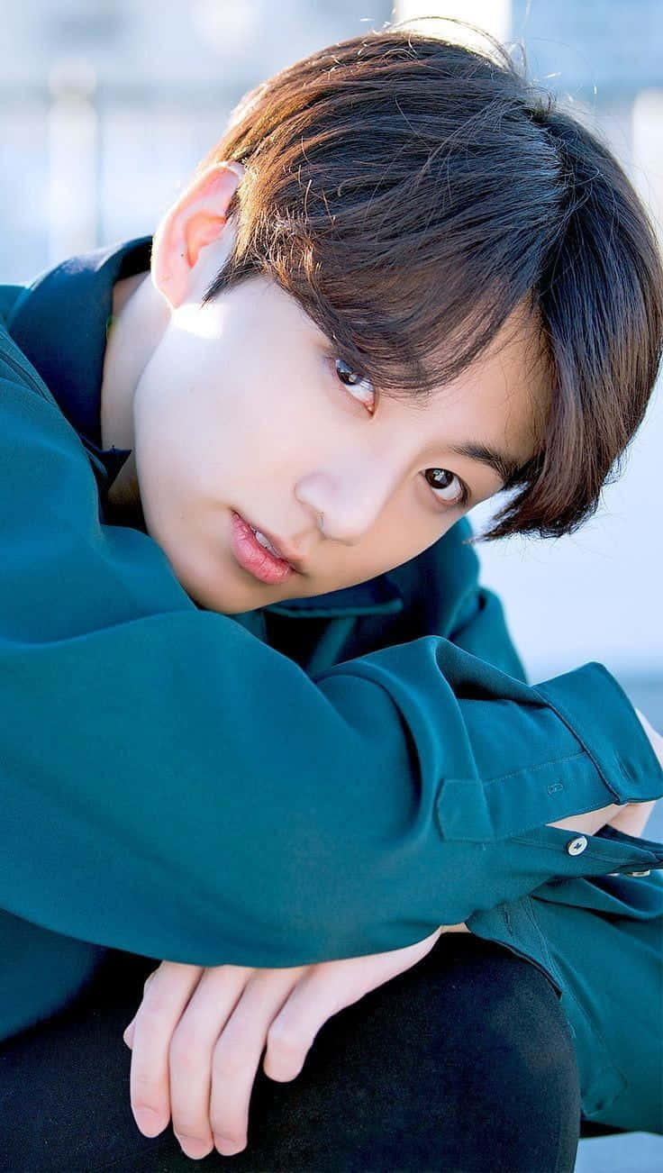 Jungkook 桌布 壁紙 — 一位深色頭髮的年輕男子穿著青綠色襯衫，在明亮模糊的背景前向前傾身。