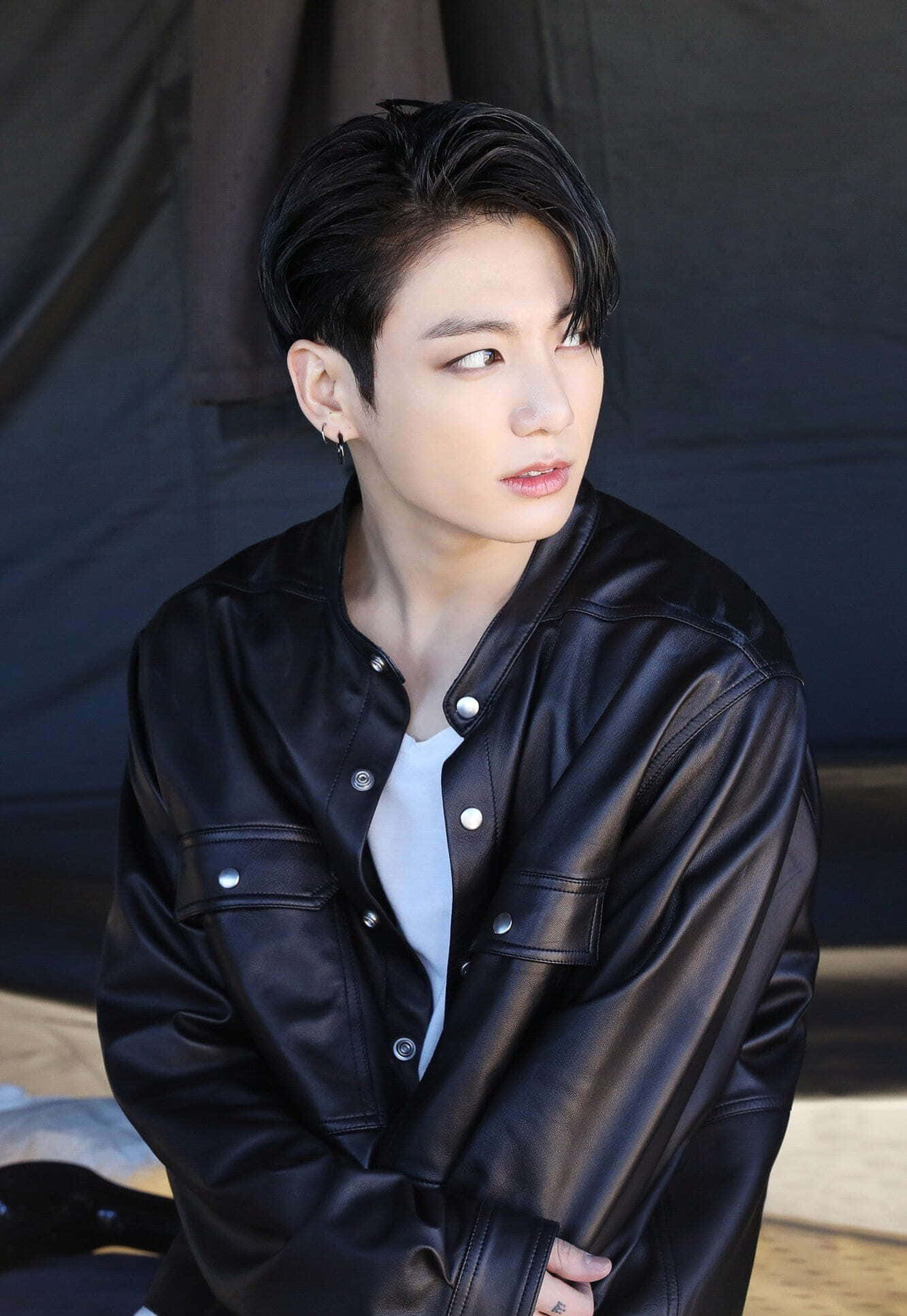Jungkook 桌布 壁紙 — 一位深色頭髮的年輕男子穿著黑色皮夾克內搭白色T恤，在深色背景下側目而視。
