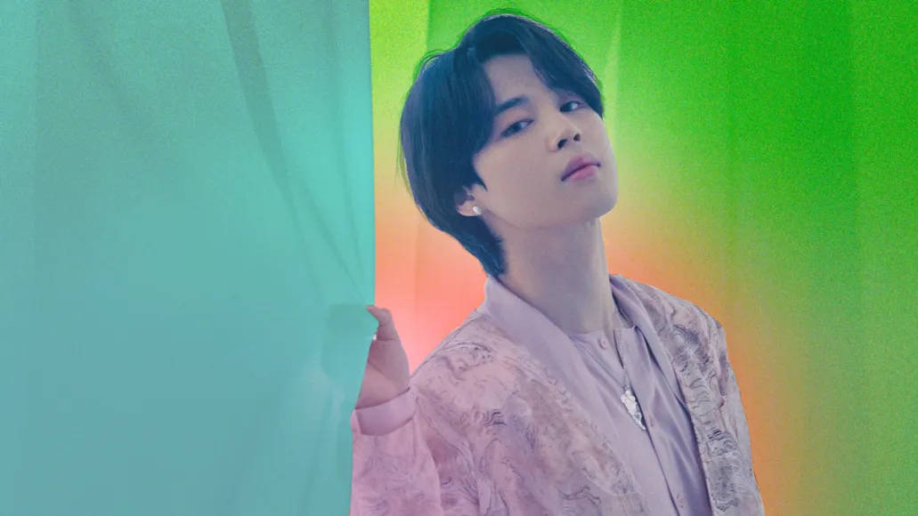 Bts Jimin 桌布 壁紙 — 一位深色頭髮的年輕男子身穿粉紅色圖案襯衫，站在藍綠色窗簾旁，背景是充滿活力的綠色和橙色漸變。