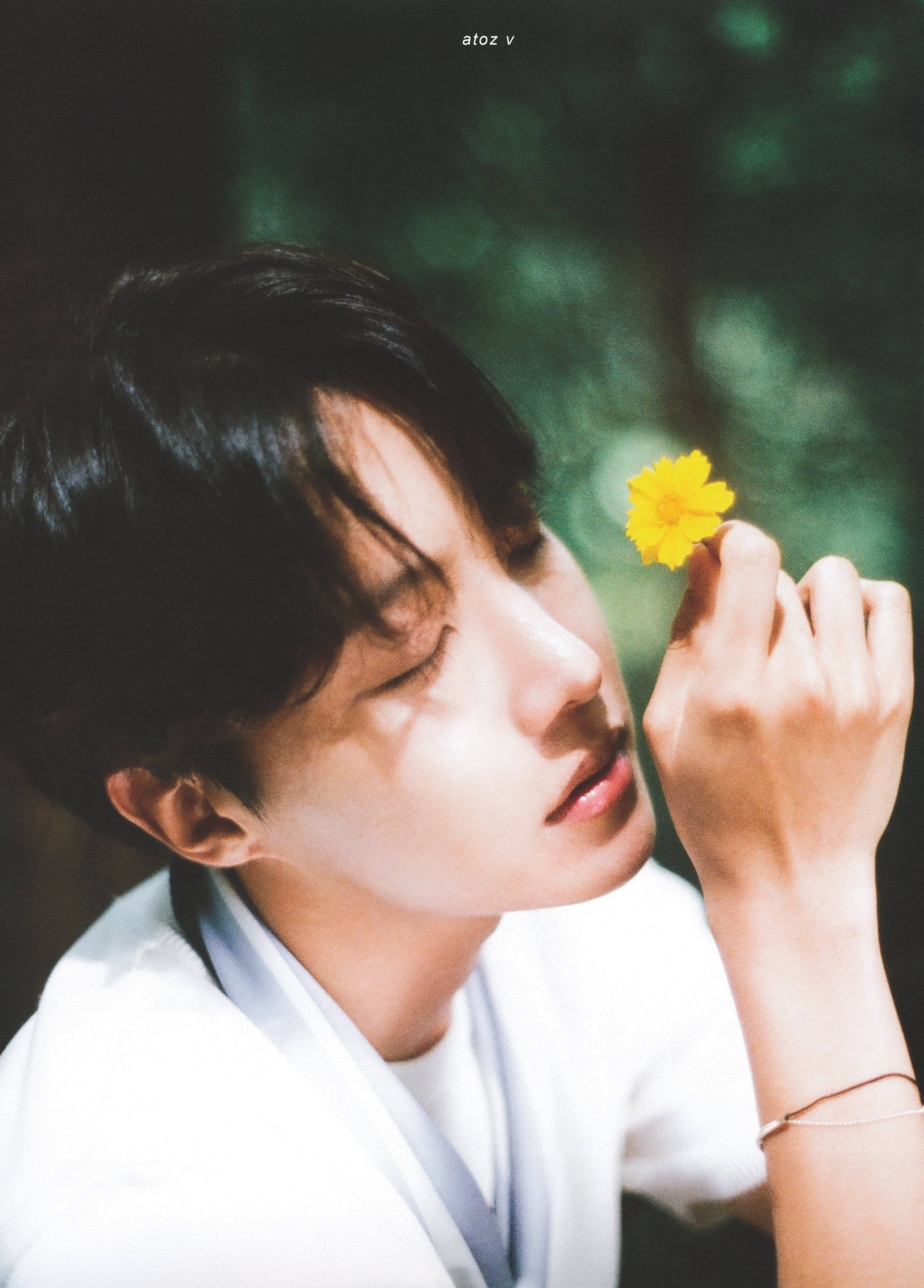 jhope 桌布 壁紙 — 一位深色頭髮的年輕男子輕輕地將一朵亮黃色的花朵放在閉上的眼睛附近，背景是柔和的綠色模糊背景。
