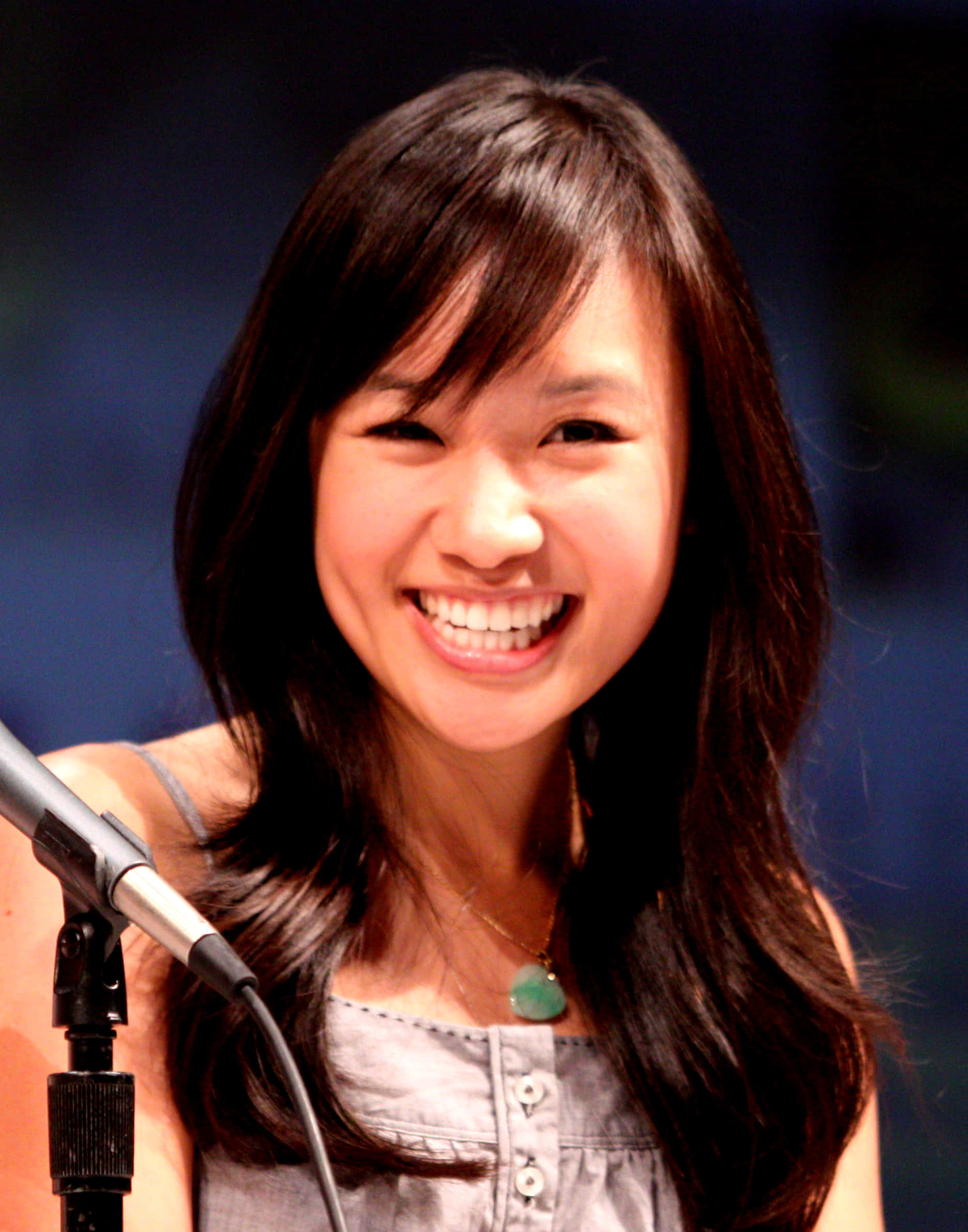 Ellen Wong 桌布 壁紙 — 一位深色頭髮的快樂女性，在模糊的藍色背景前對著麥克風燦爛微笑說話。