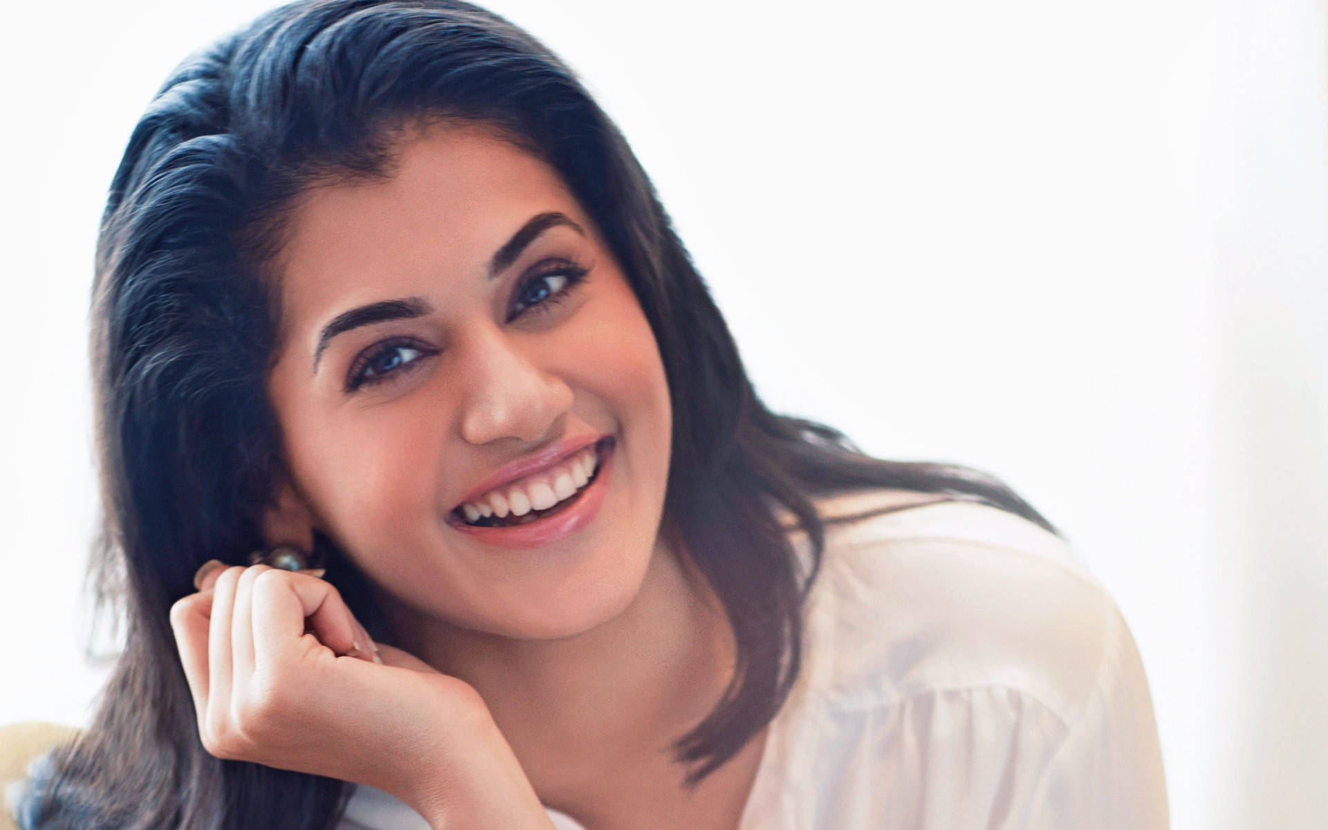 Taapsee Pannu 桌布 壁紙 — 一位深色頭髮的燦爛女性，在明亮的白色背景前，將下巴擱在手上，溫暖地微笑。