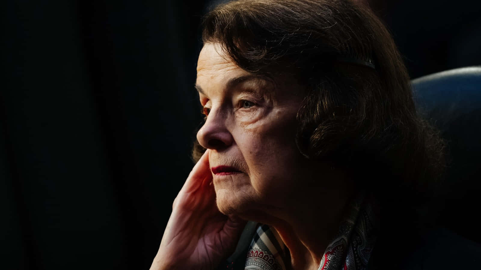 Dianne Feinstein 桌布 壁紙 — 一位深色頭髮的老年女性在昏暗的環境中將手放在臉頰上，她的臉被柔和的光線照亮。