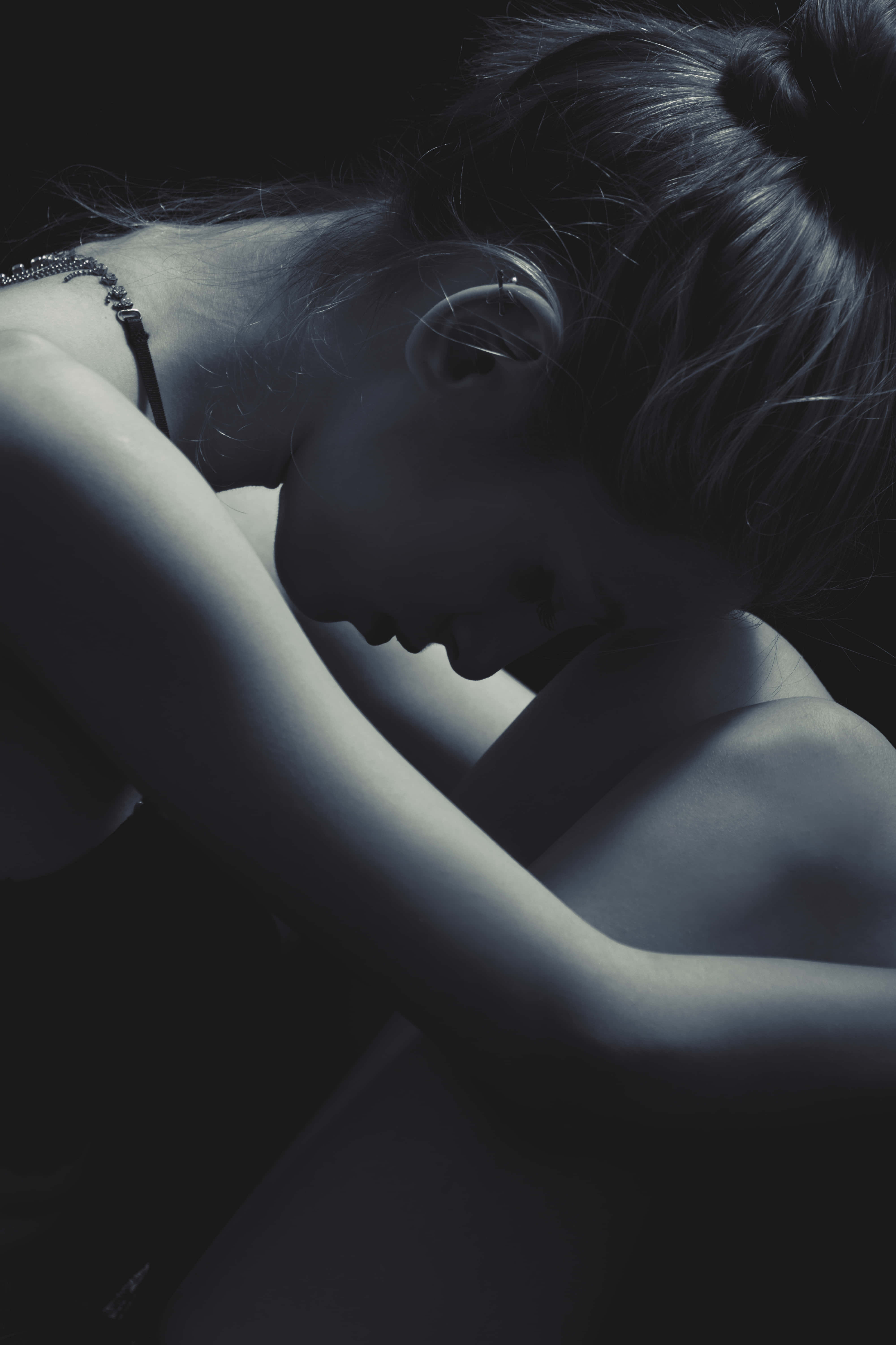 憂鬱美學 桌布 壁紙 — 一位深色頭髮盤成髮髻的女性，在昏暗、憂鬱的藍調肖像中將頭靠在手臂上。