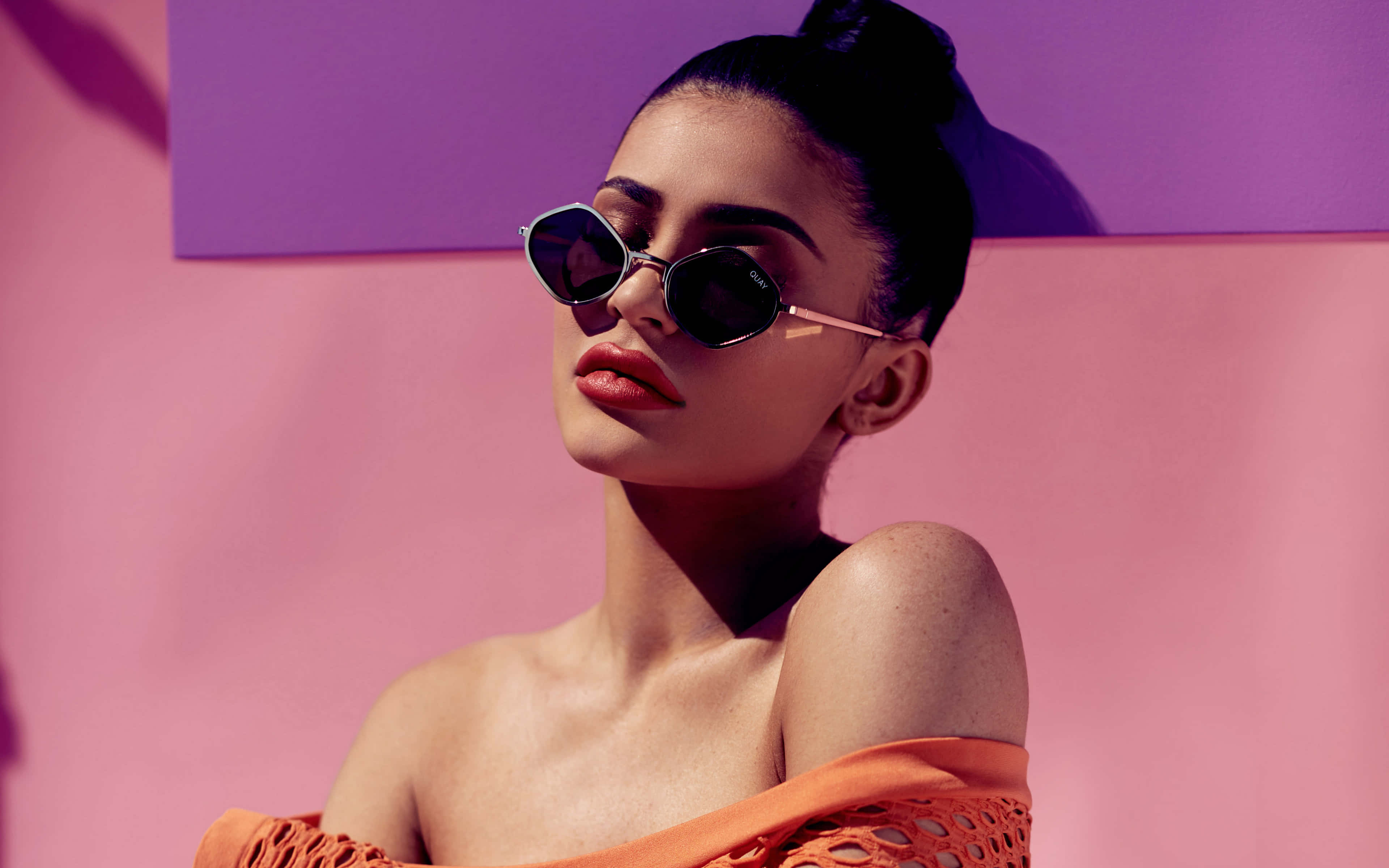 Kylie Jenner 4K 桌布 壁紙 — 一位深色頭髮盤成髮髻的女性，戴著圓形太陽眼鏡和橙色露肩上衣，背景是鮮豔的粉紅色和紫色。