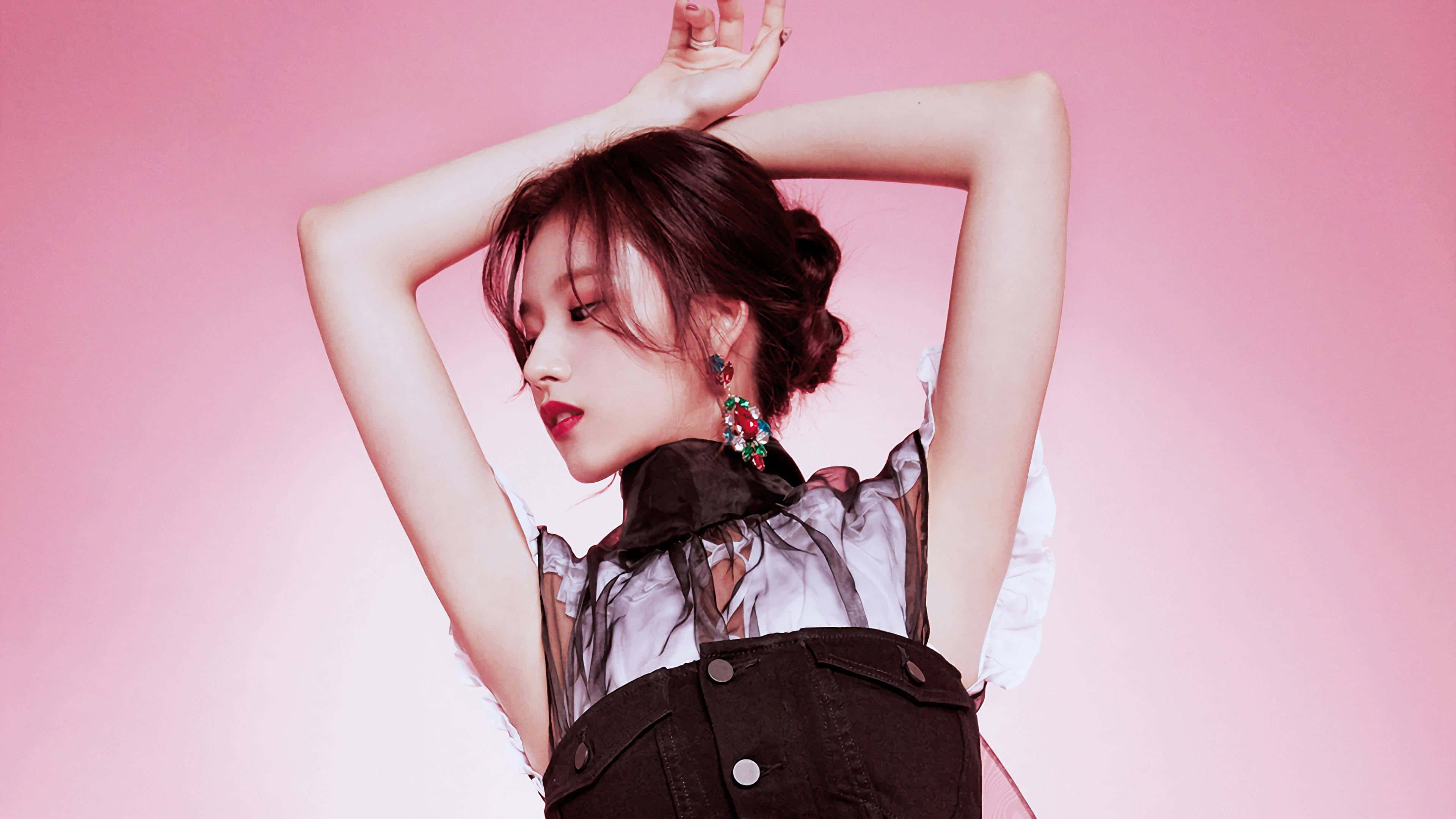 twice 桌布 壁紙 — 一位深色頭髮盤成髮髻的年輕女性，在柔和的粉紅色工作室背景前優雅地擺姿勢，雙臂舉起，穿著透明襯衫和深色吊帶褲。