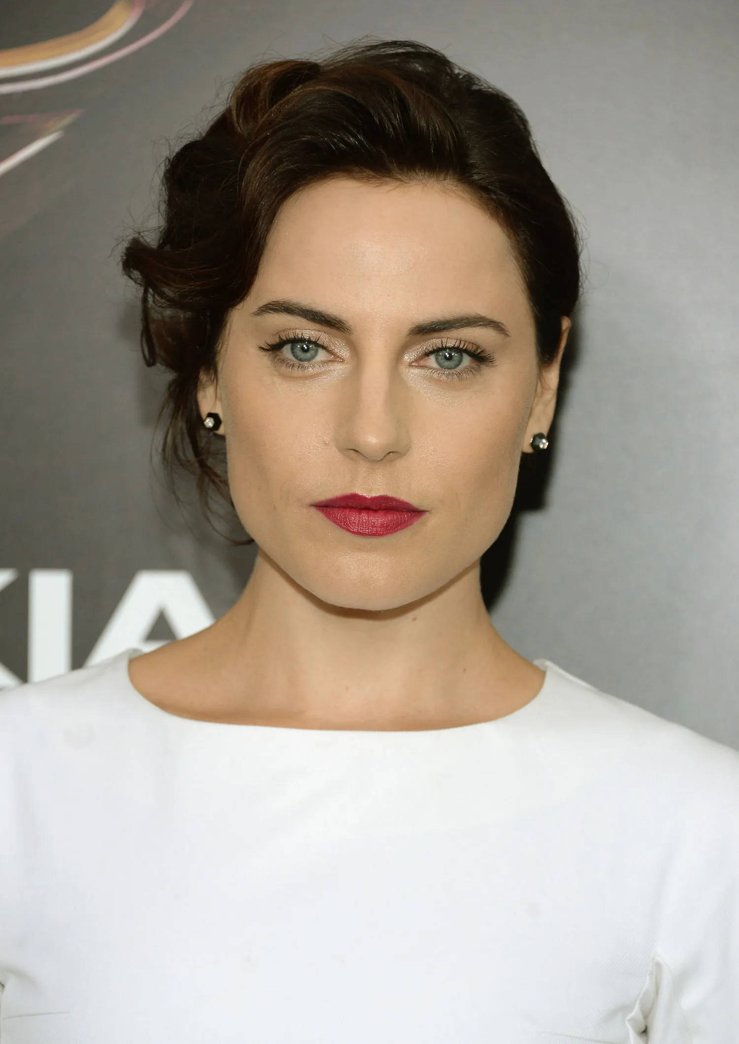 Antje Traue 桌布 壁紙 — 一位深色頭髮盤起、擁有引人注目的藍色眼睛和鮮豔紅唇的女性特寫肖像，背景為灰色。