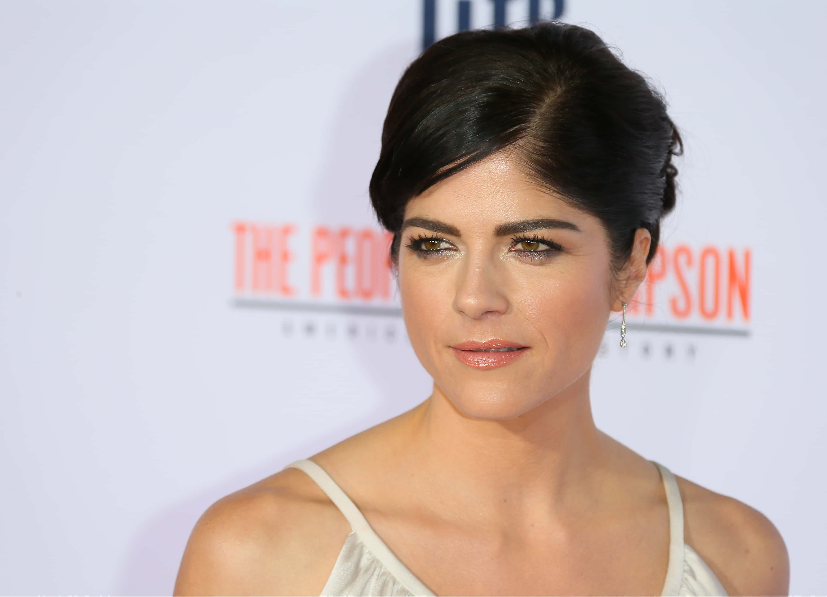 Selma Blair 桌布 壁紙 — 一位深色頭髮盤起的女士在紅地毯活動中穿著白色連衣裙和優雅的耳環。