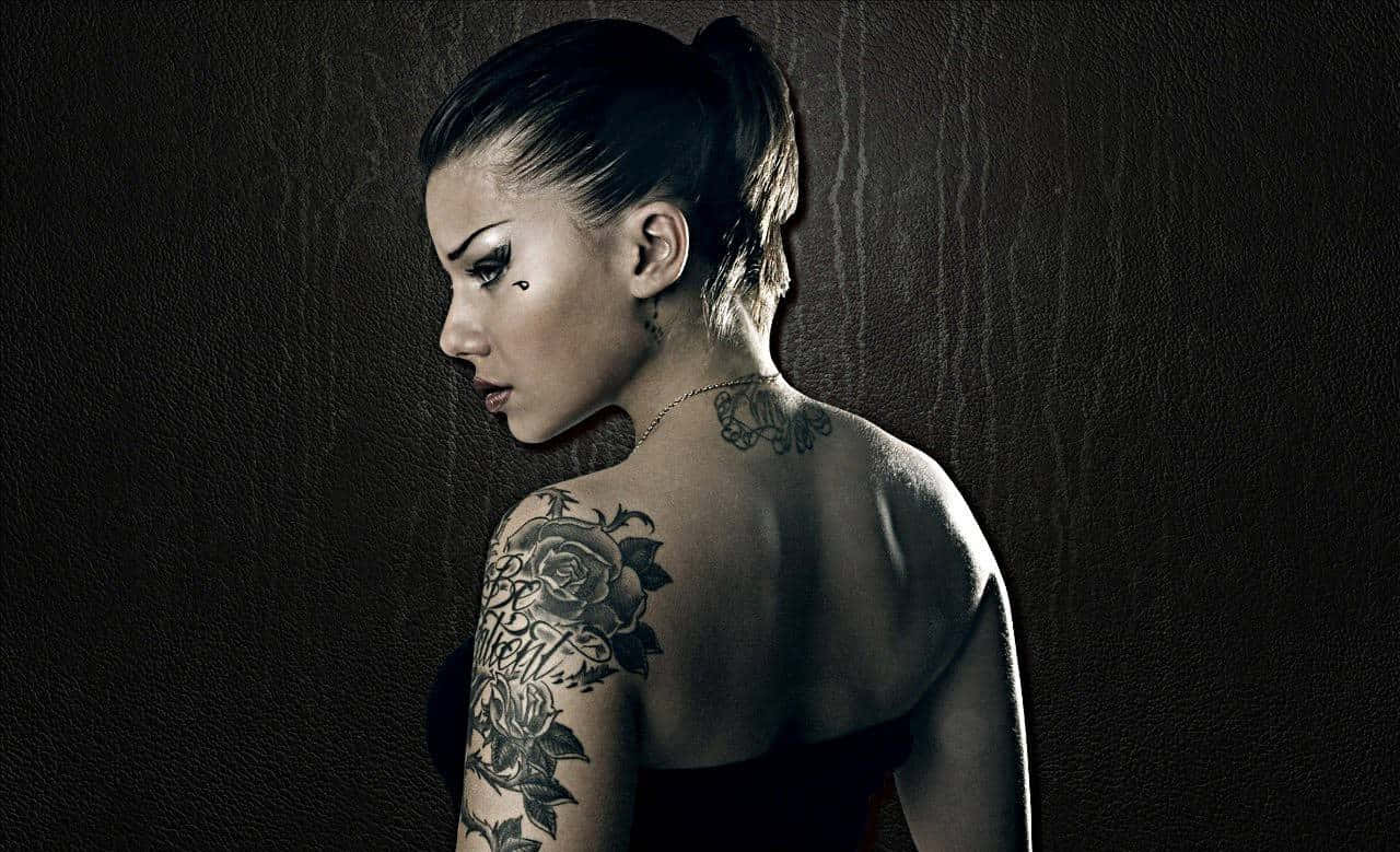 Tattoo 桌布 壁紙 — 一位深色頭髮紮成馬尾、肩膀和頸部有刺青的女性，在紋理深色牆壁前向下凝視。
