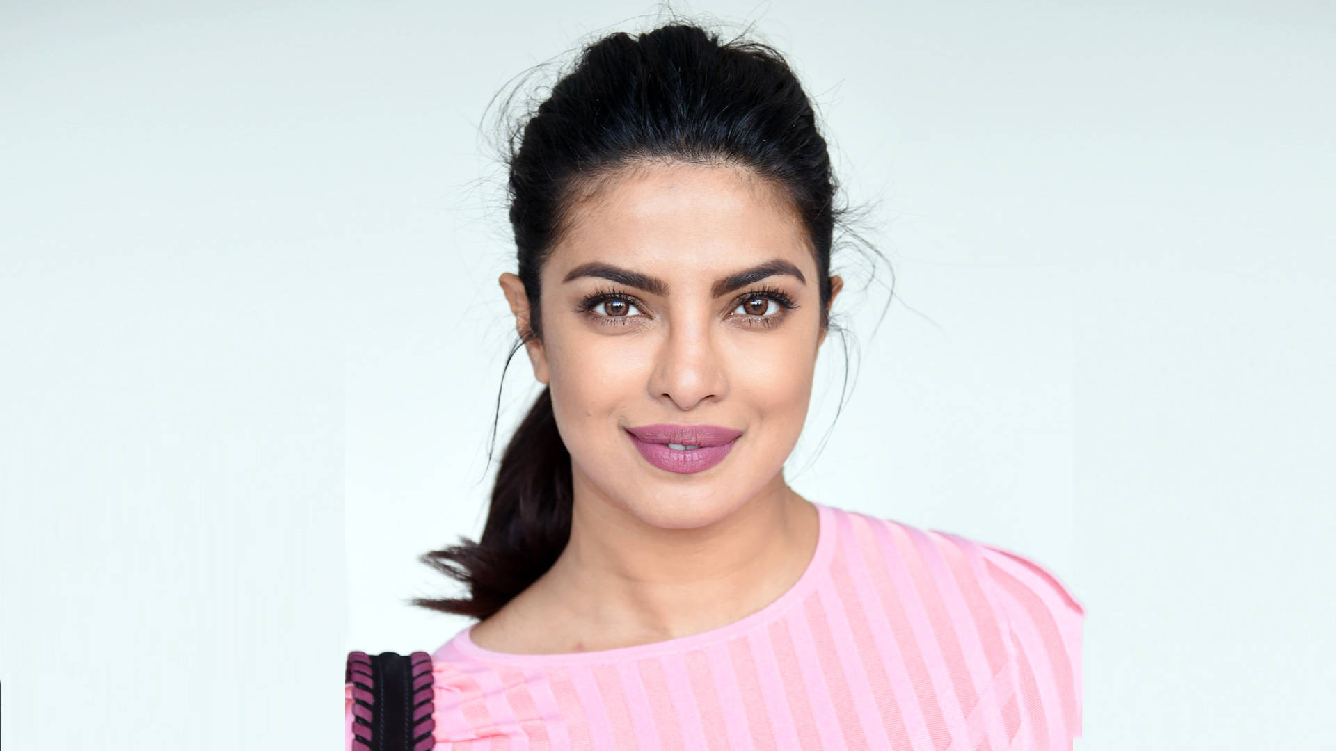 Priyanka 桌布 壁紙 — 一位深色頭髮紮成馬尾的女性，在純白色背景前穿著粉紅色條紋上衣，溫柔地微笑。
