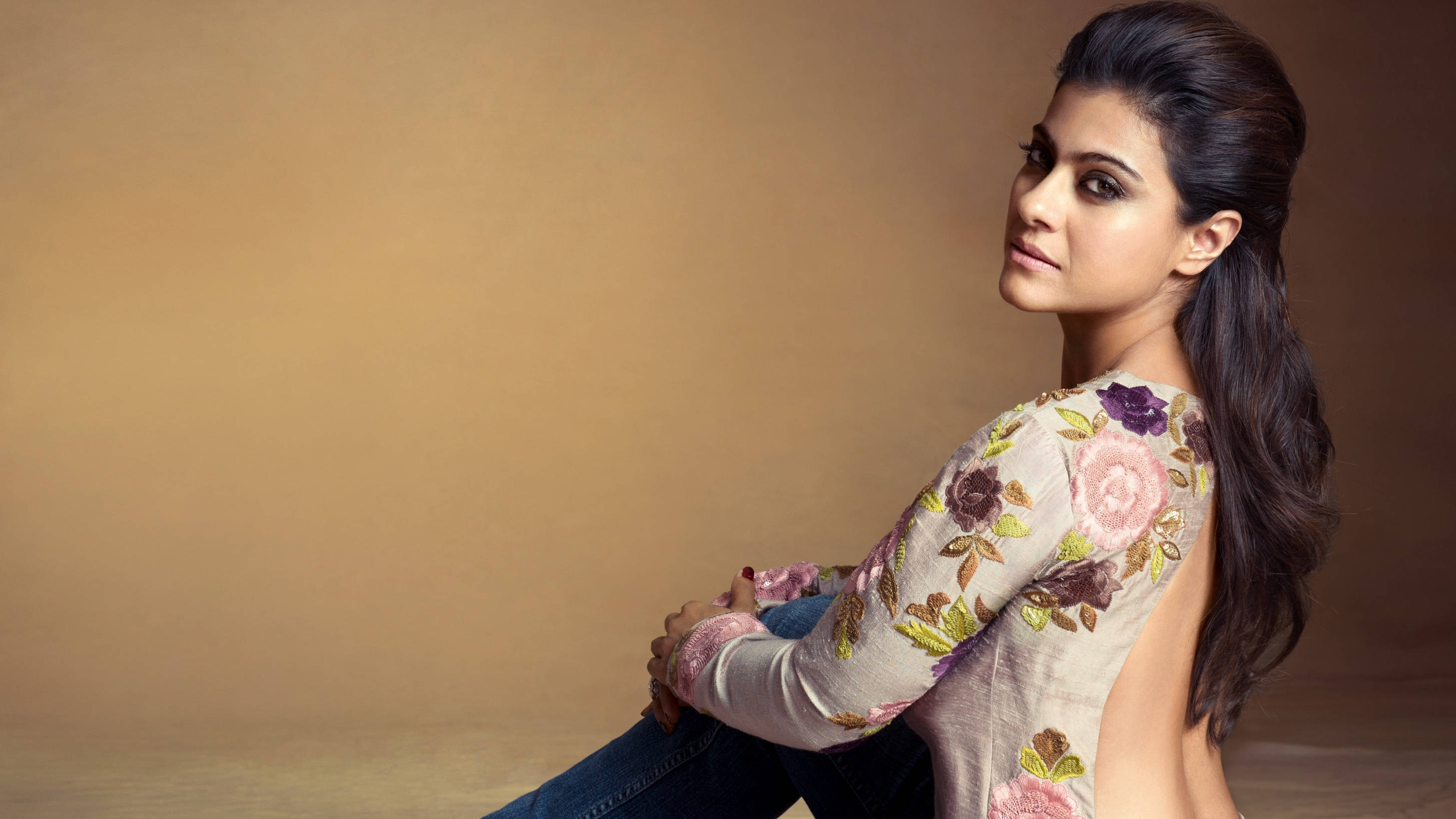 Kajol 桌布 壁紙 — 一位深色頭髮紮馬尾的女性稍微轉身坐著，穿著花卉刺繡上衣，背景是溫暖的米色工作室。