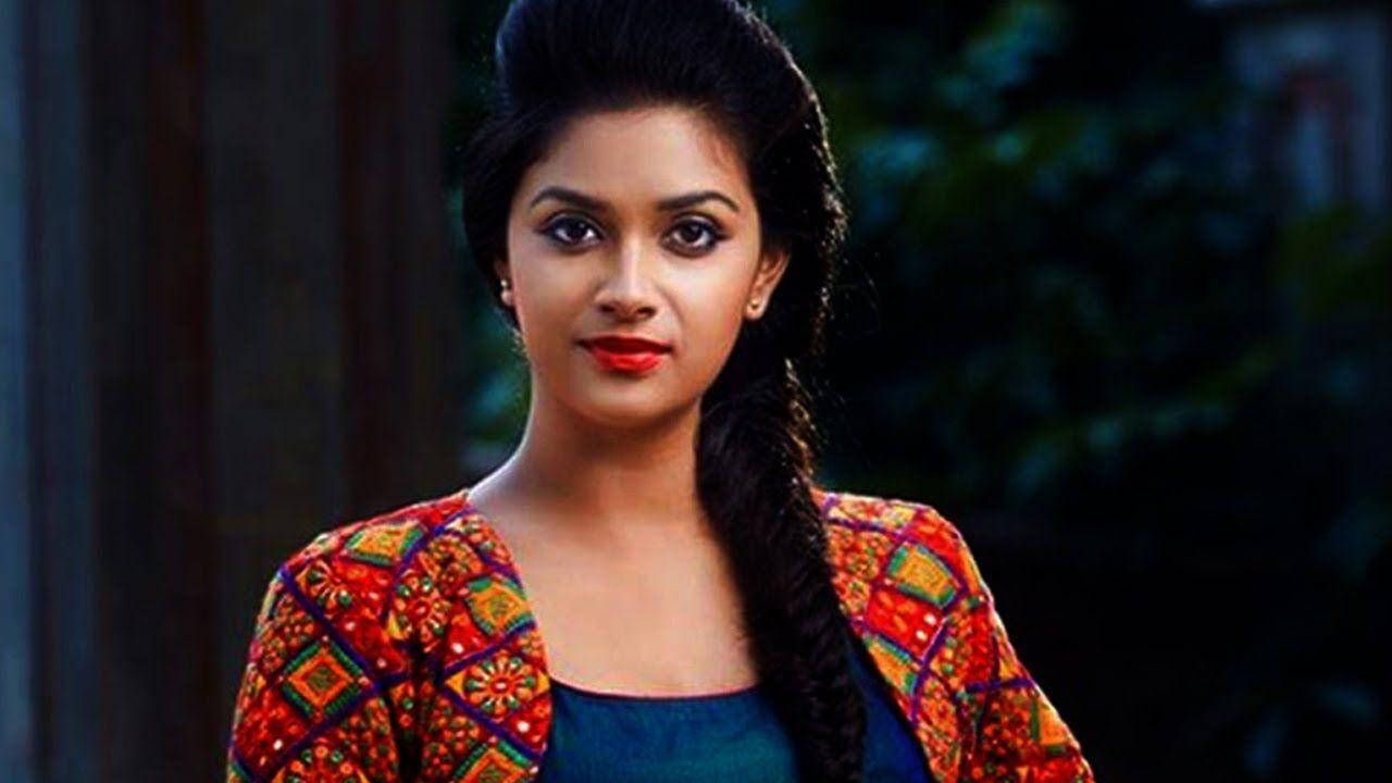Keerthi Suresh HD 桌布 壁紙 — 一位深色頭髮編成辮子的女性，在模糊的綠色背景前，穿著一件彩色圖案外套，內搭青綠色上衣。