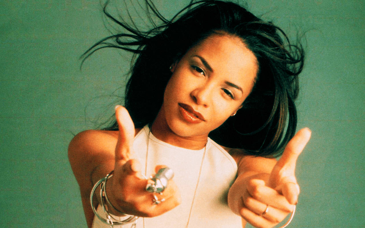 aaliyah 桌布 壁紙 — 一位深色頭髮隨風飄動的女性，在青綠色背景前豎起兩個大拇指。