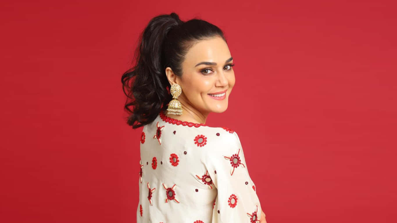 Preity Zinta 桌布 壁紙 — 一位深色頭髮高馬尾的女性，在純紅色背景上回頭微笑，穿著白色花卉圖案上衣，戴著金色耳環。