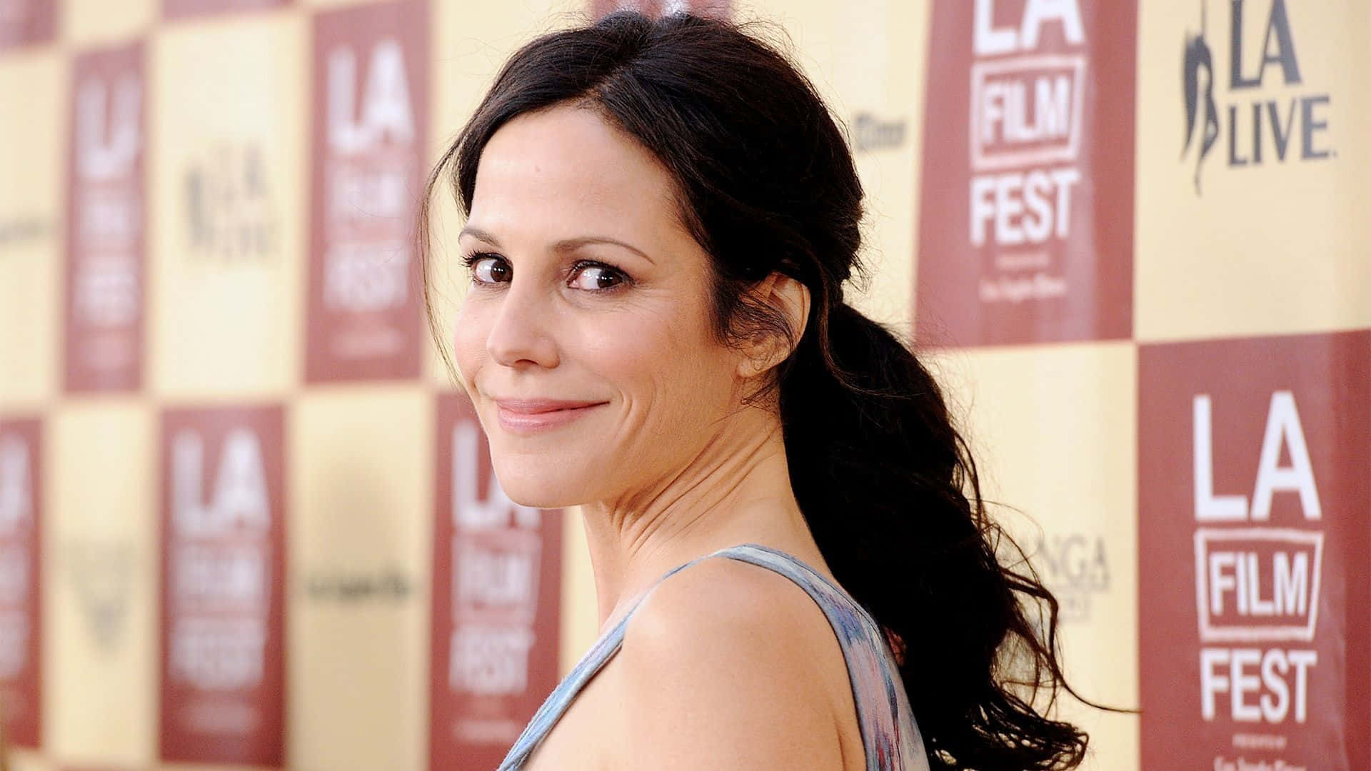 mary louise parker 桌布 壁紙 — 一位深色馬尾辮的女性在紅色和米色的LA Film Fest背景前回頭微笑。