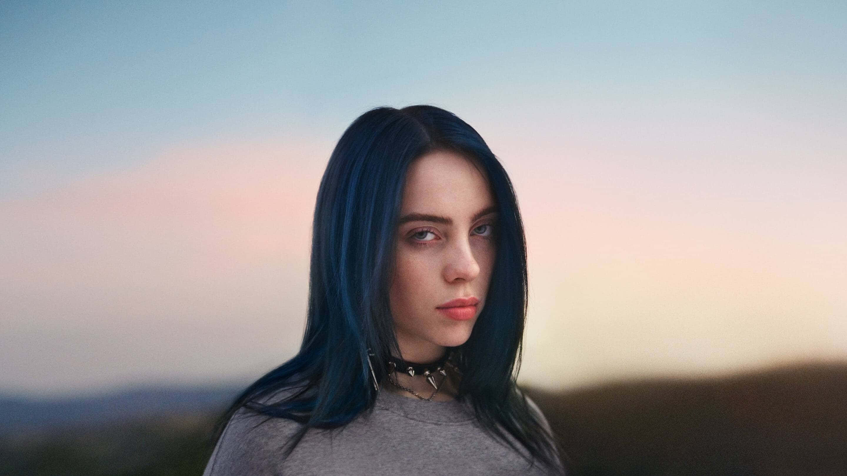 Billie Eilish Laptop 桌布 壁紙 — 一位深藍色頭髮的年輕女性，在柔和的粉彩日落天空背景下直視鏡頭。