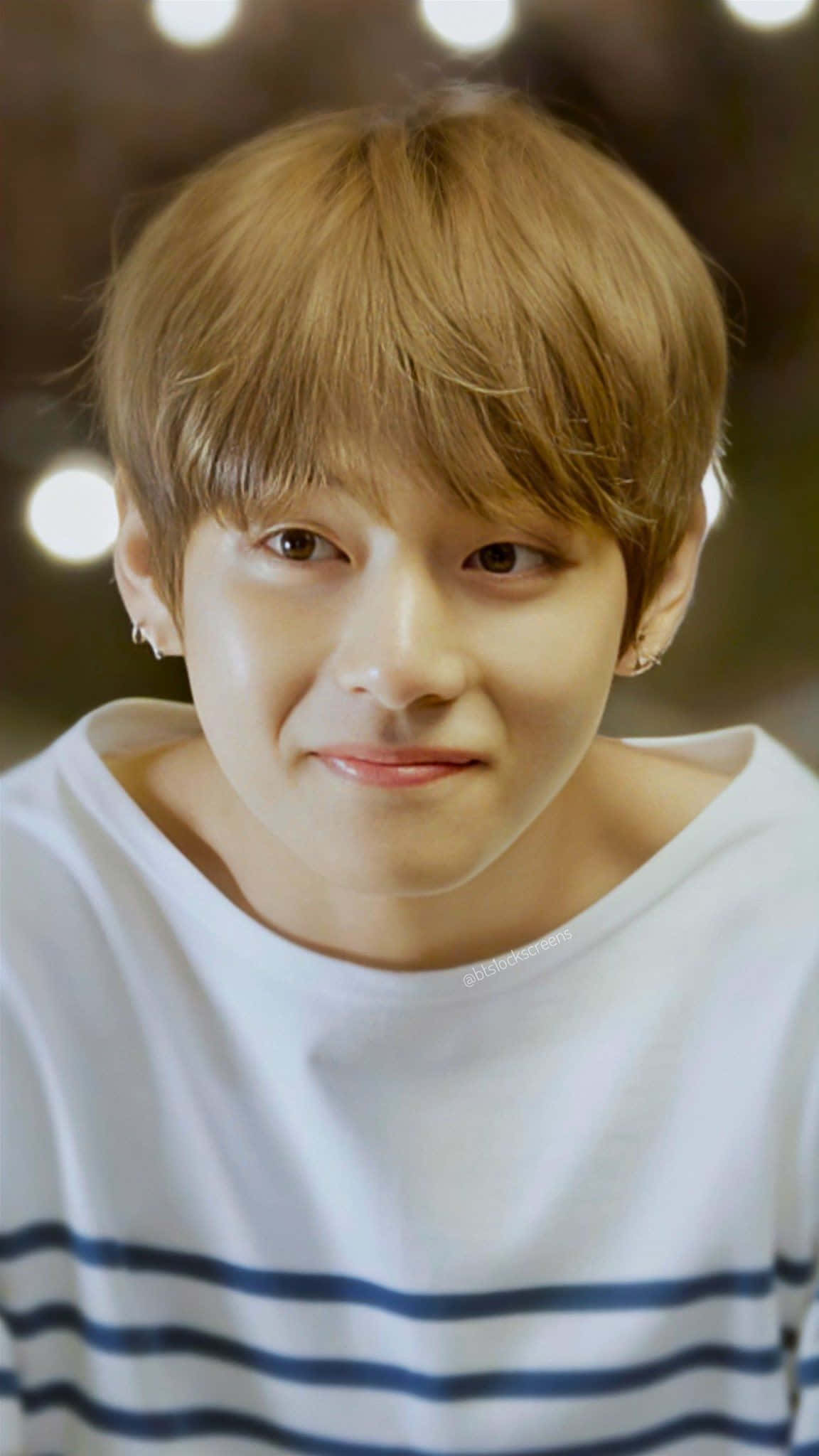 Taehyung Cute 桌布 壁紙 — 一位淺棕色頭髮、帶著溫柔微笑的年輕男子，穿著白色條紋襯衫，背景是柔和模糊的溫暖燈光。