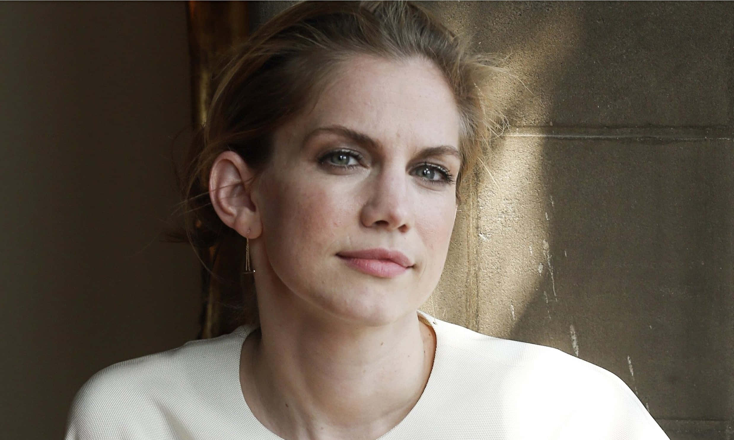anna chlumsky 桌布 壁紙 — 一位淺棕色頭髮、藍色眼睛的女性，在溫暖陽光照耀的紋理石牆前若有所思地凝視。