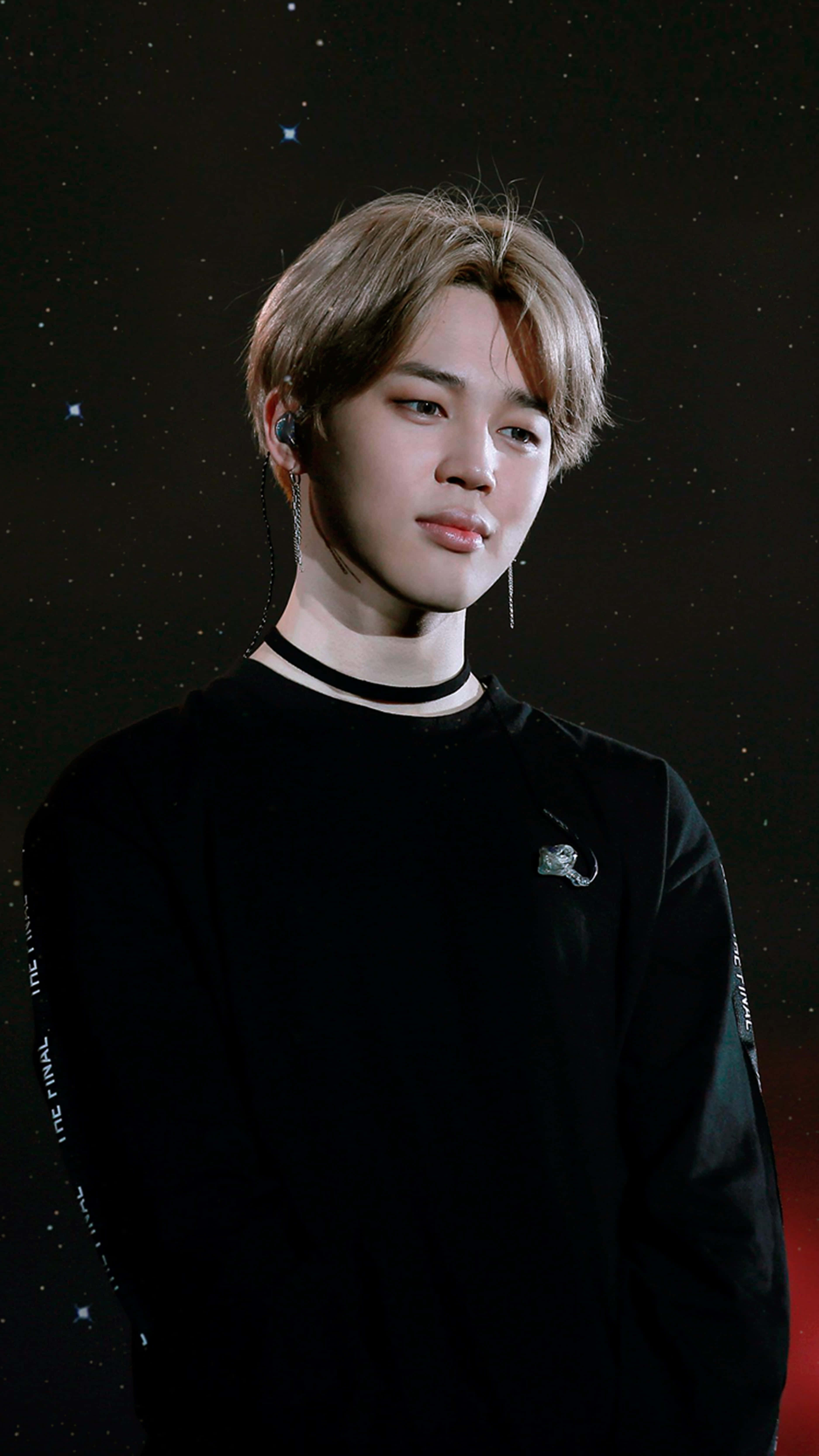 BTS 桌布 壁紙 — 一位淺棕色頭髮、身穿黑色服裝的年輕男子，站在繁星點點的夜空背景下，帶有微妙的紅色光芒。