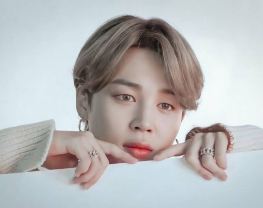 BTS 智旻 桌布 壁紙 — 一位淺棕色頭髮和紅唇的年輕男子，將下巴擱在白色表面上，戴著戒指和針織毛衣，背景是柔和的白色。