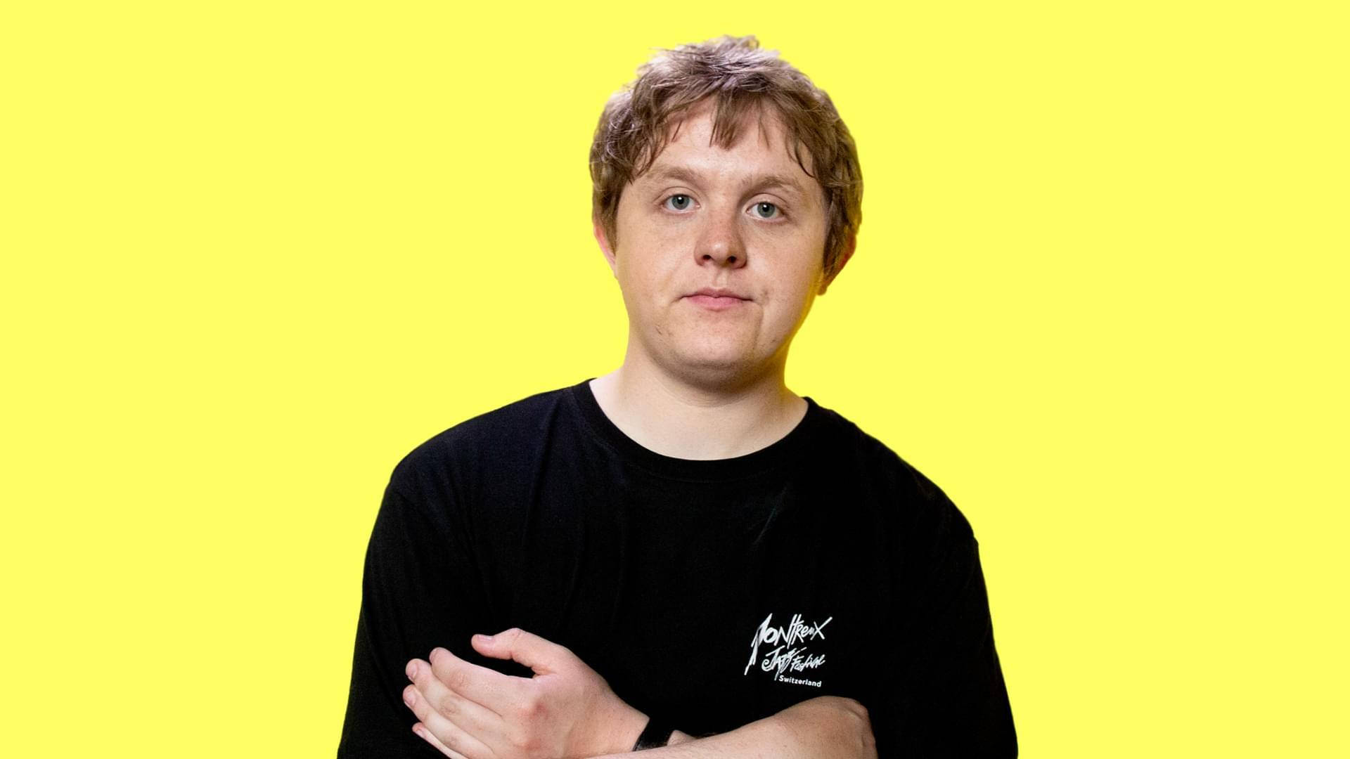 Lewis Capaldi 桌布 壁紙 — 一位淺棕色頭髮的年輕男子自信地交叉雙臂，站在鮮黃色背景前，身穿黑色T恤。