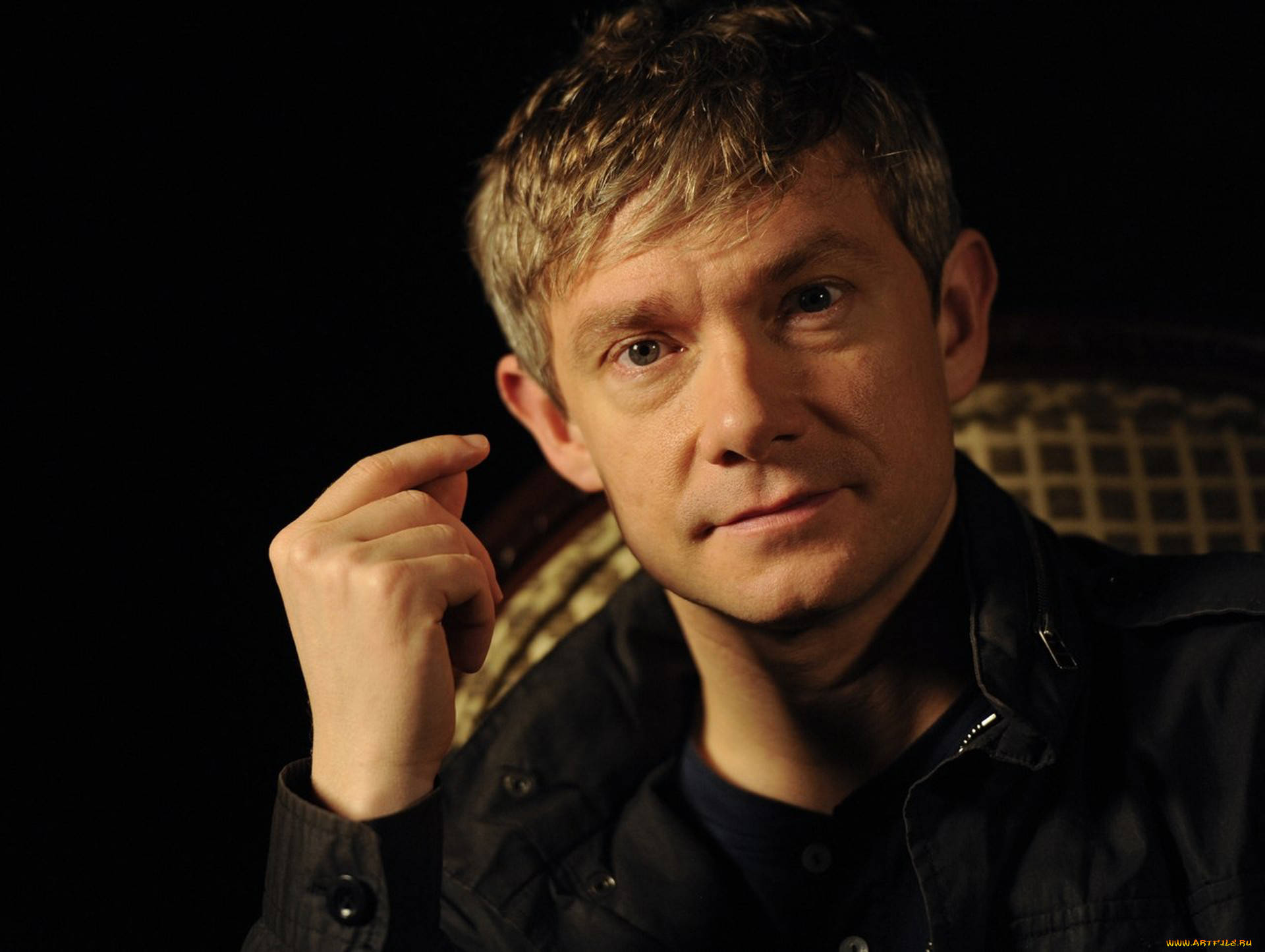 Martin Freeman 桌布 壁紙 — 一位淺色頭髮、眼神銳利的男士坐在昏暗的房間裡，手靠近臉部，身穿深色外套。