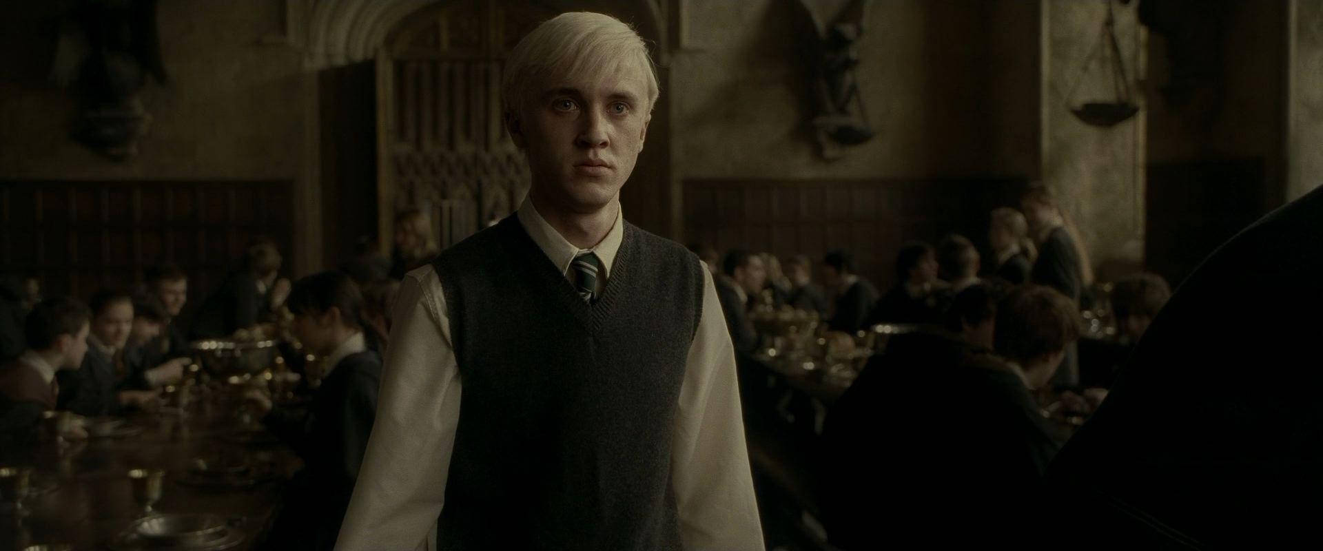 draco 桌布 壁紙 — 一位淺色頭髮的年輕男子穿著校服，莊嚴地站在一個光線昏暗、擠滿學生的飯廳裡。