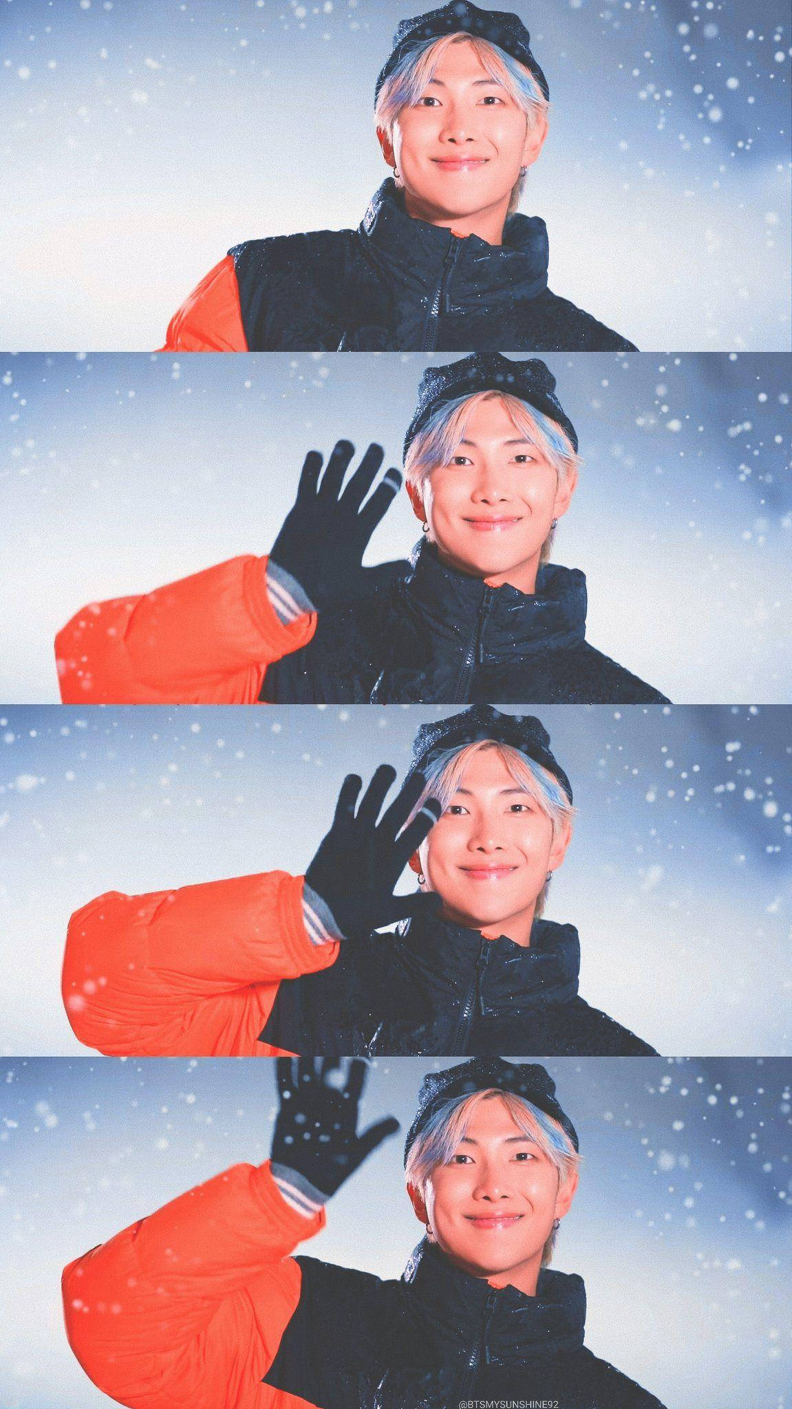 BTS 桌布 壁紙 — 一位淺藍色頭髮的年輕人在飄雪中穿著黑橙色冬季外套愉快地揮手。