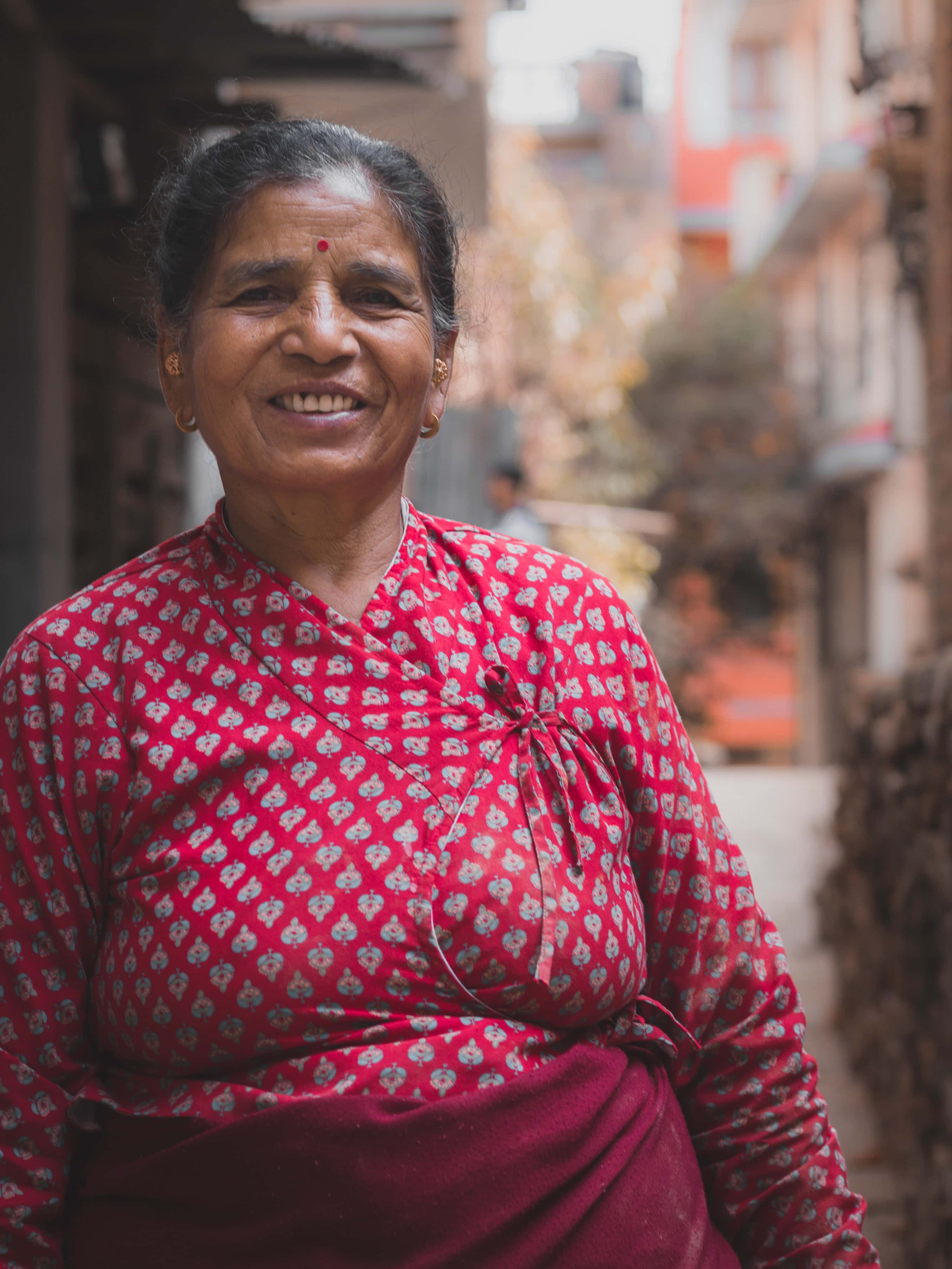 Indian 桌布 壁紙 — 一位溫暖微笑的女子穿著紅色圖案襯衫，站在兩旁有建築物的陽光照射狹窄小巷中。