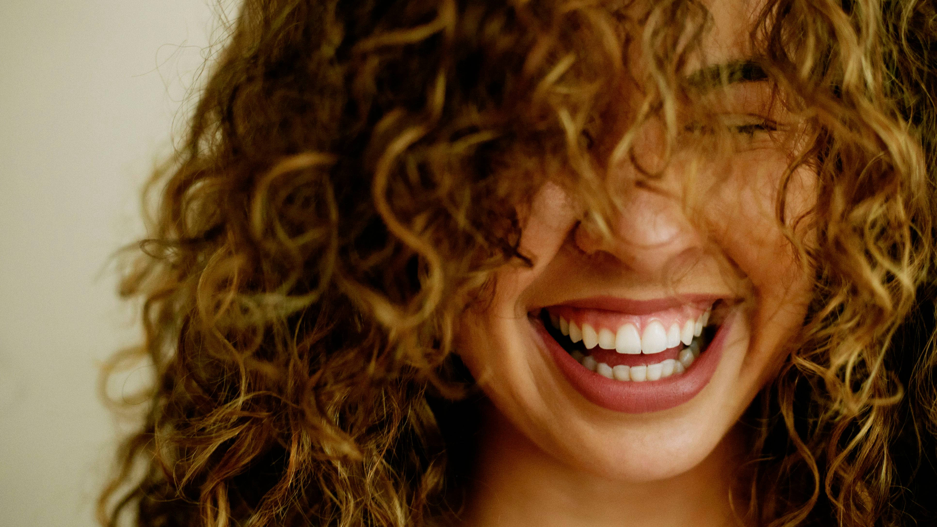 Smile 桌布 壁紙 — 一位滿懷喜悅、擁有一頭豐盈鬈髮的女性開懷大笑，她燦爛的笑容在溫暖的金色調中露出潔白的牙齒。