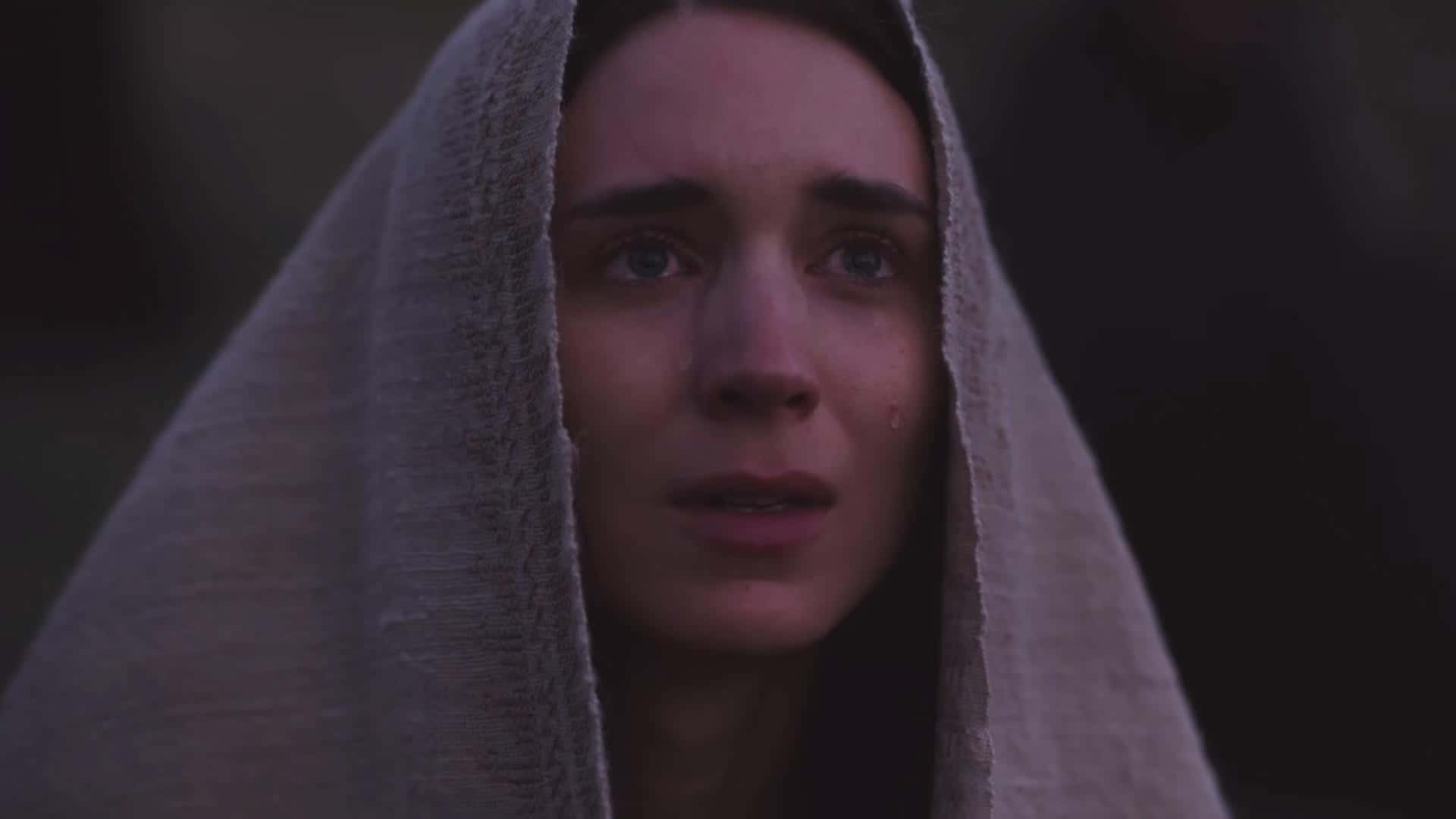 Mary Magdalene 桌布 壁紙 — 一位滿臉淚痕、睜大眼睛的女性，在昏暗光線下從有紋理的灰色連帽下探出頭來。