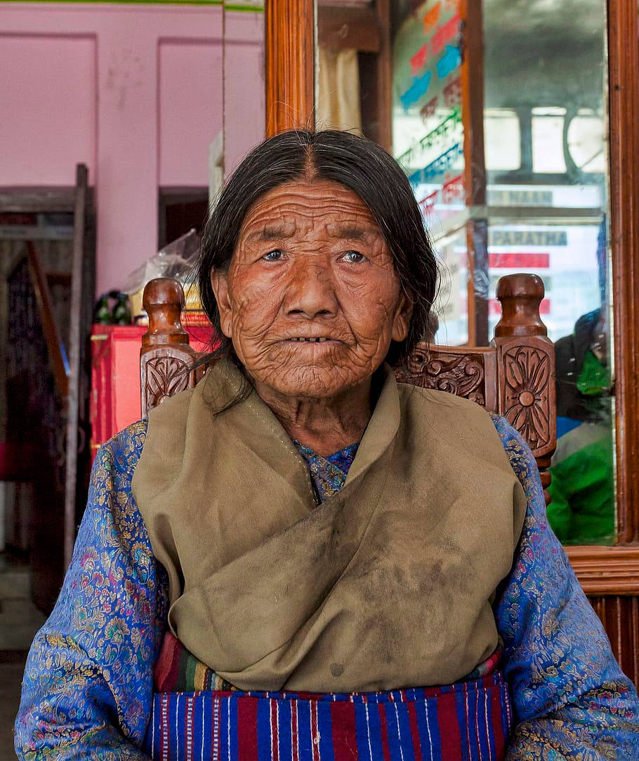 Woman 桌布 壁紙 — 一位滿臉皺紋、眼神銳利的老婦人，身穿傳統藍色圖案服裝和米色圍巾，靜靜地坐在木椅上。